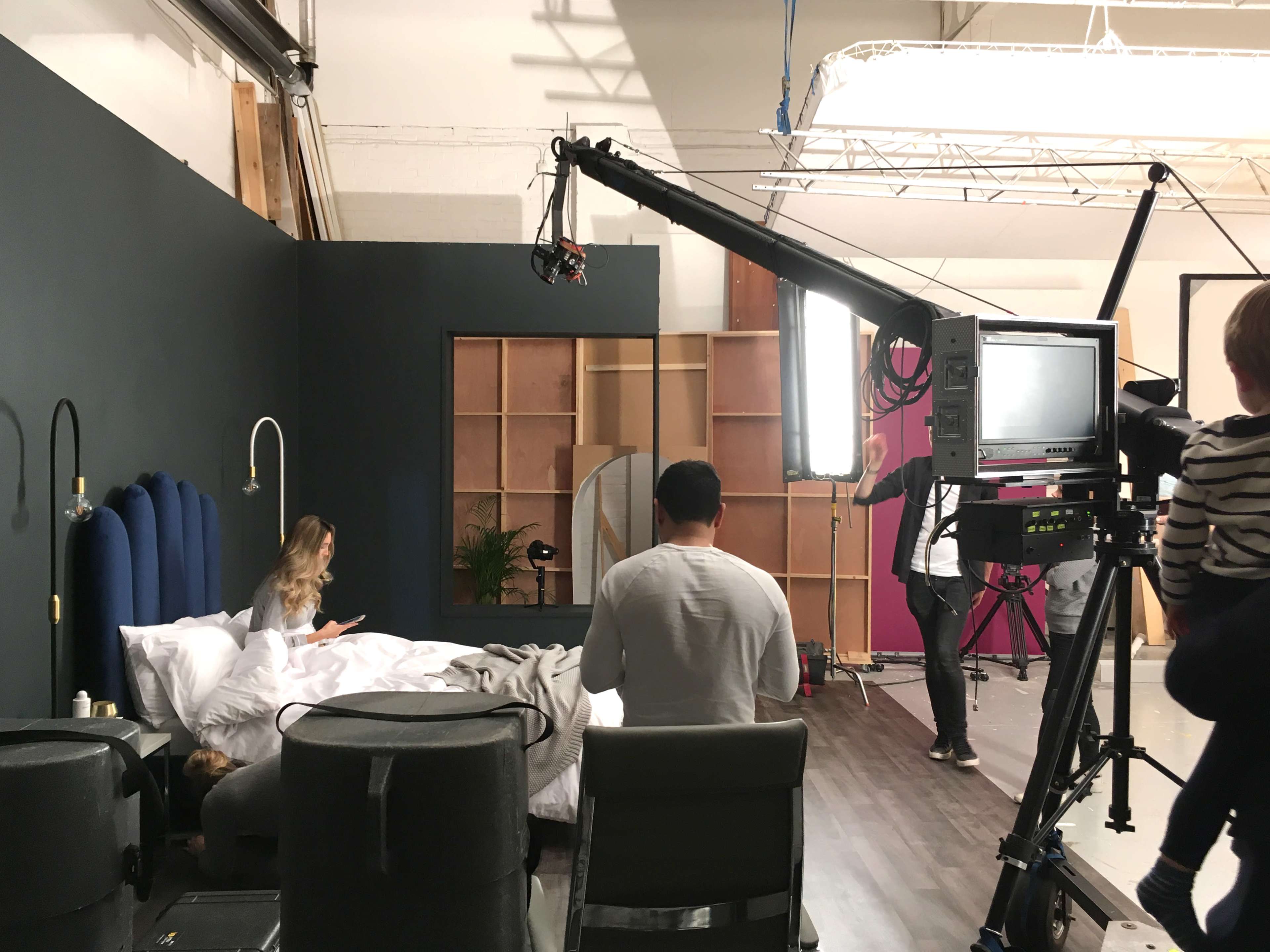 A film crew sets up a scene in a studio bedroom with a woman sitting on the bed and a camera mounted on a crane.