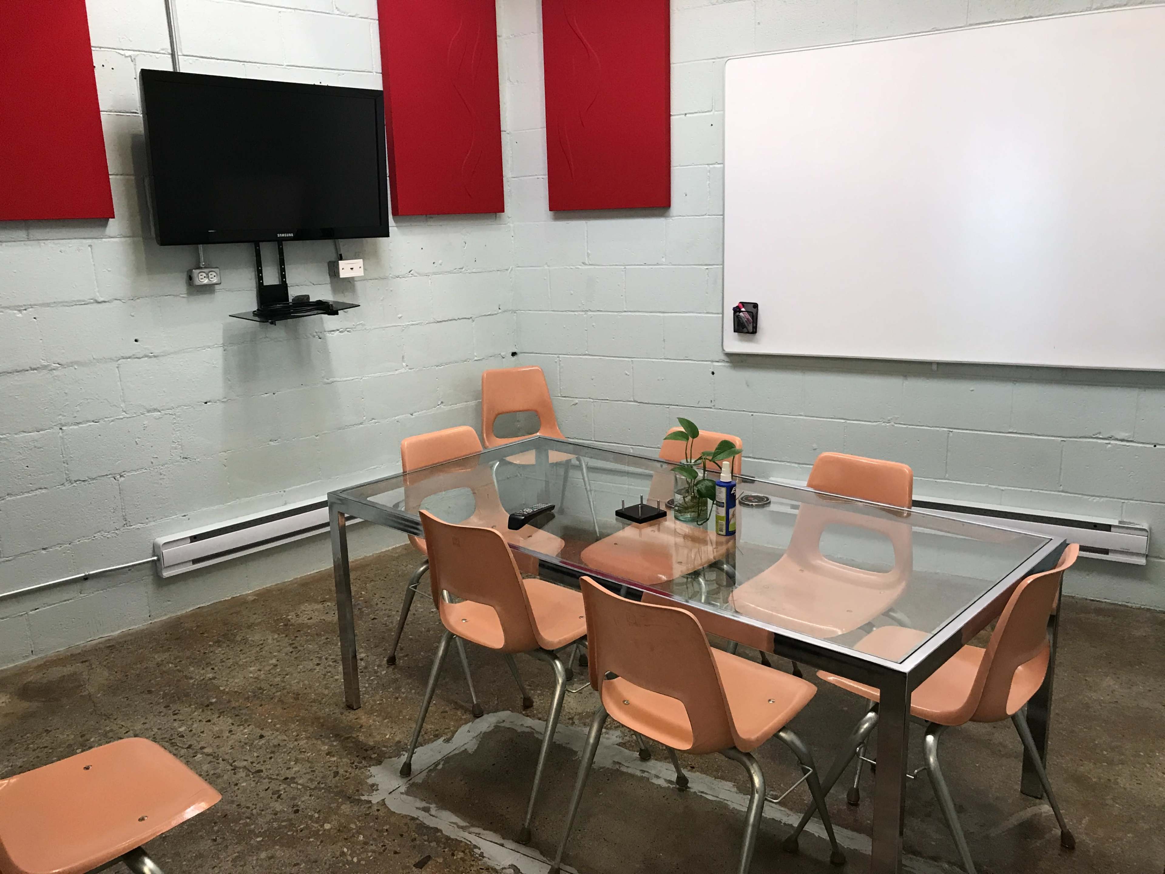 A meeting room features a glass table surrounded by eight peach-colored chairs, a wall-mounted television, and a whiteboard.