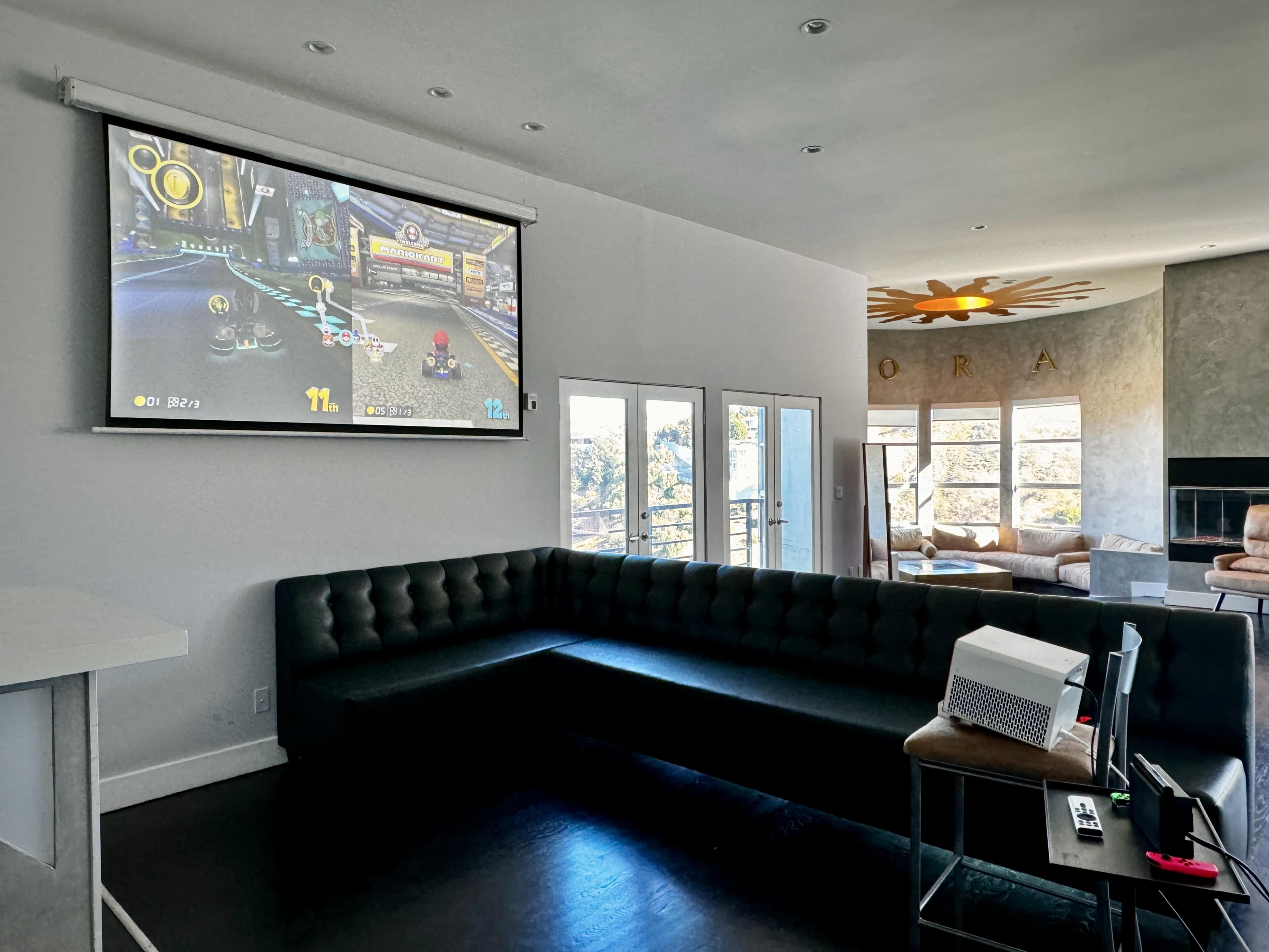 A large projection screen displays a video game in a modern living room with a sectional sofa and a view of the outdoors.