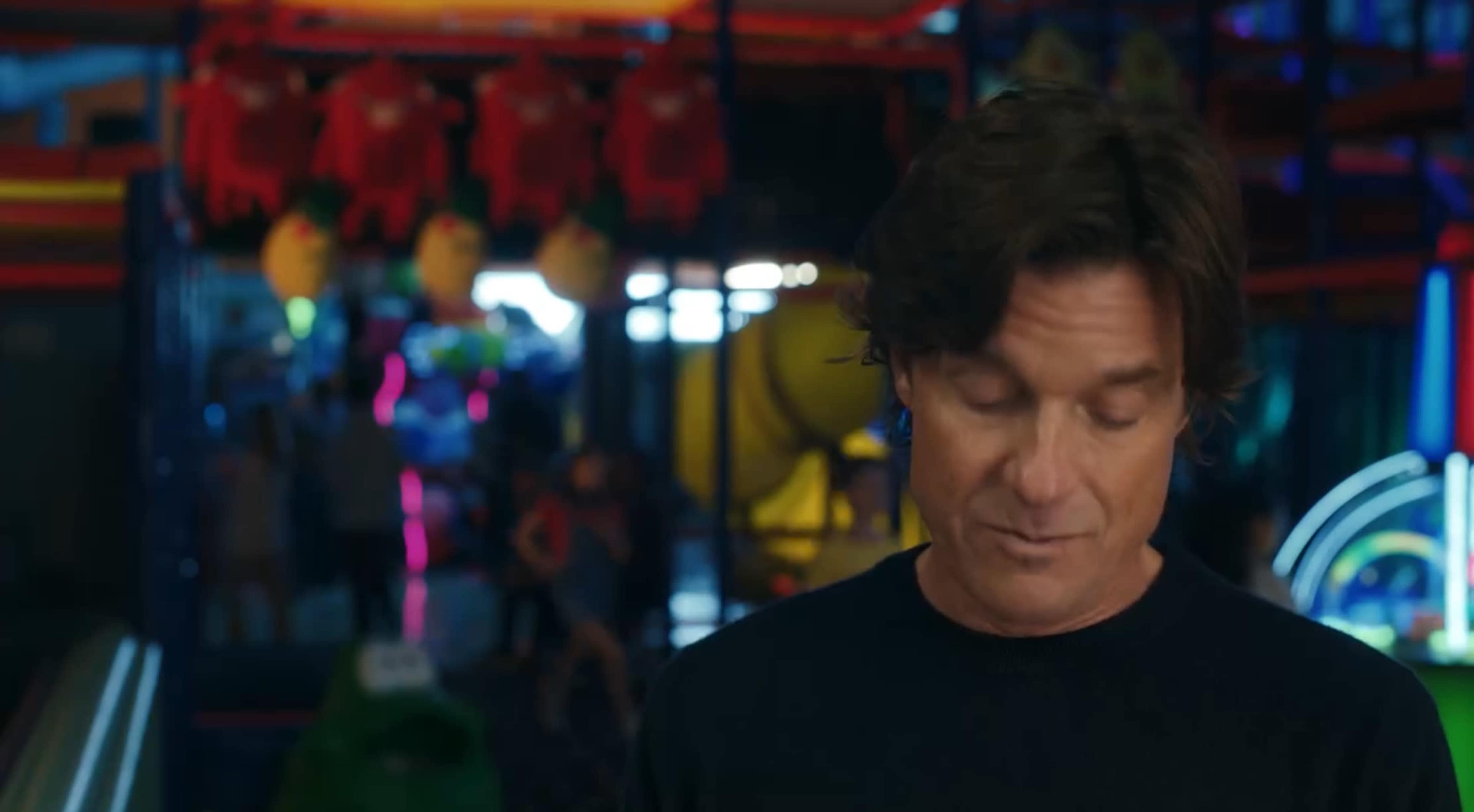 A man with dark hair stands in the foreground of an arcade filled with colorful lights and various games in the background.