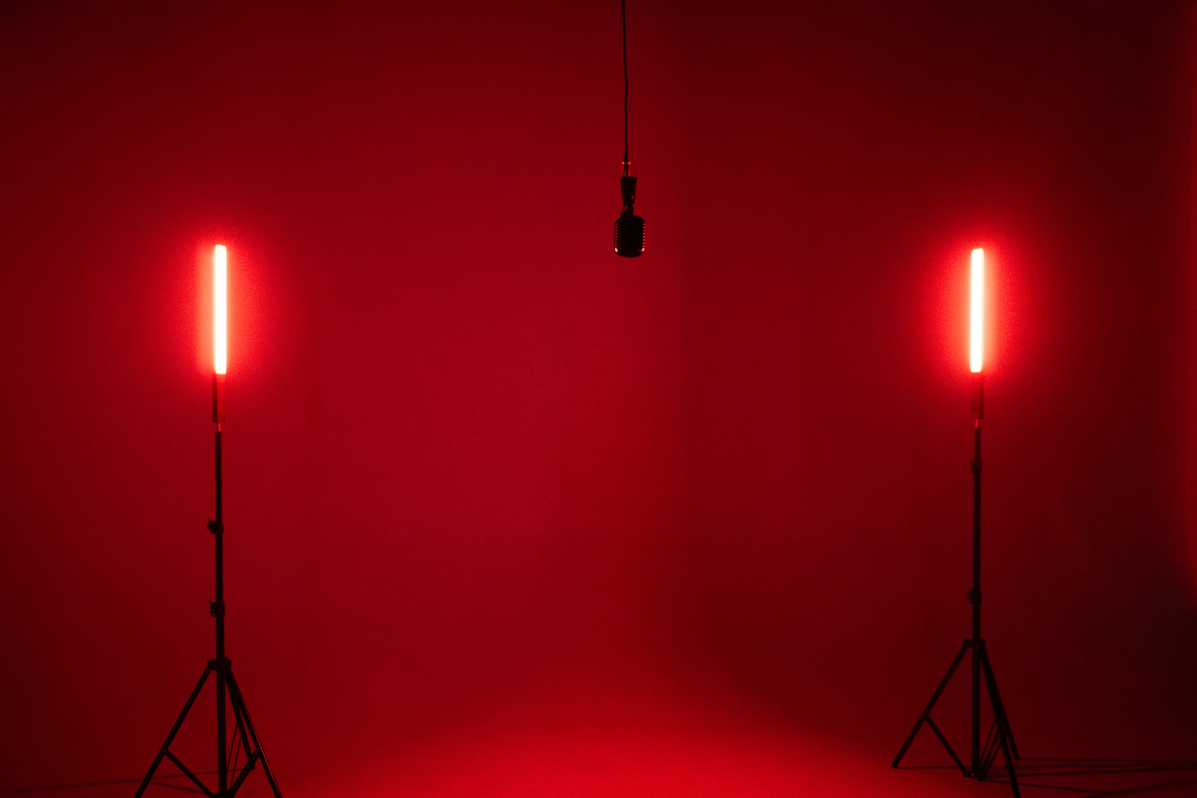 The image shows a minimalistic stage setup with a microphone hanging in the center, flanked by two red lights on stands and a deep red background.