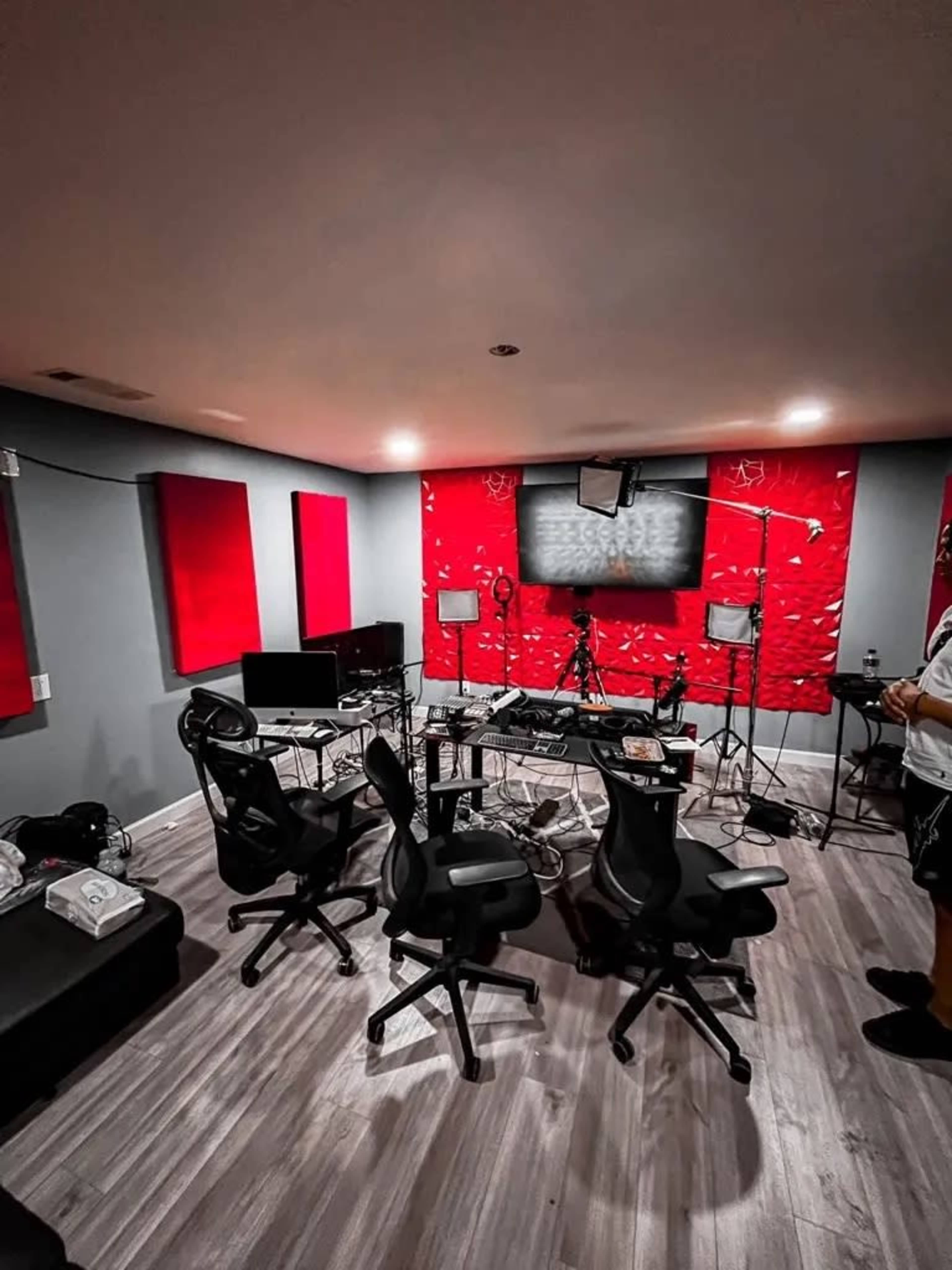 A recording studio is furnished with multiple chairs, computers, lighting equipment, and a red acoustic panel backdrop.