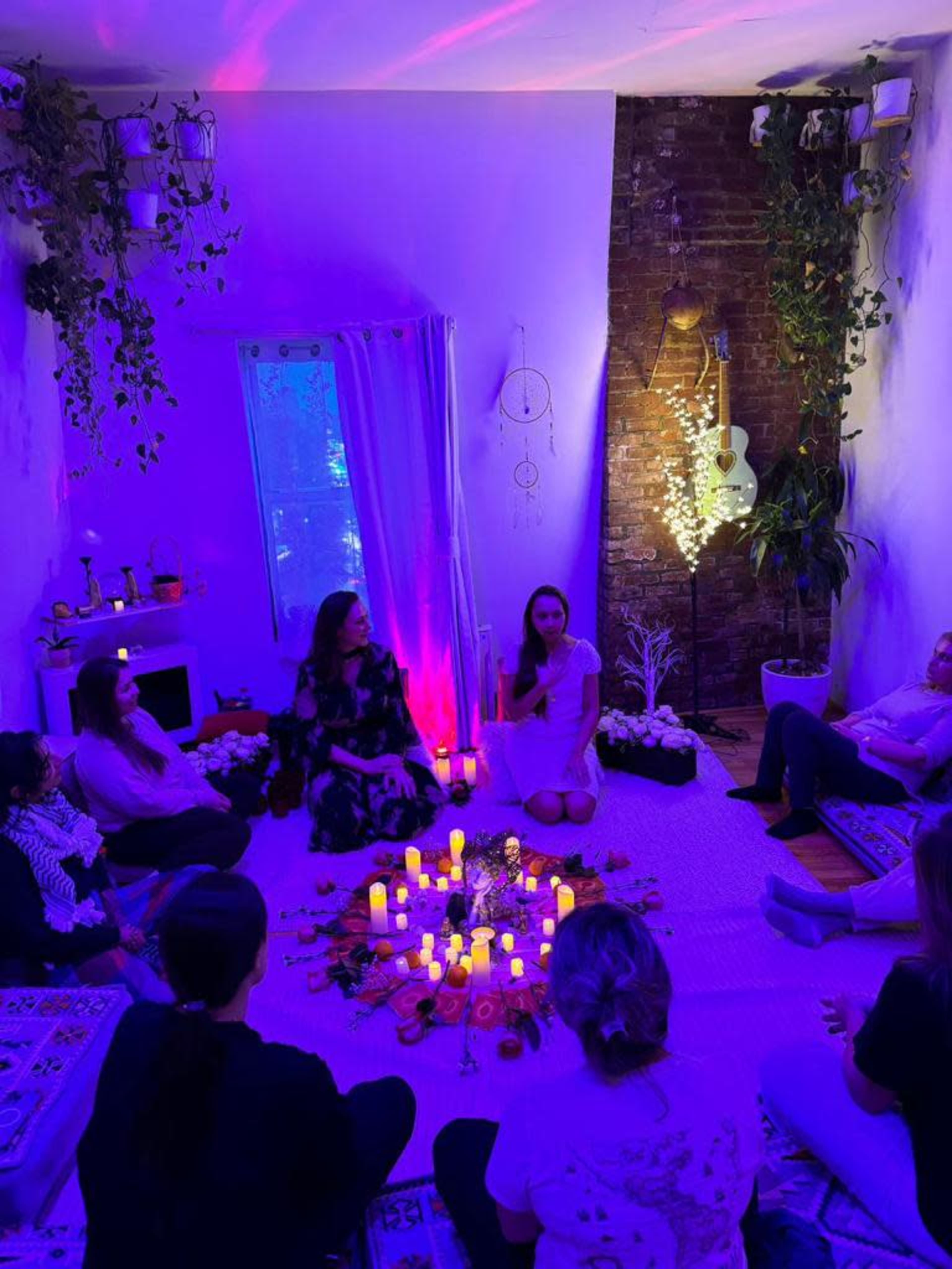 A group of people sits on the floor in a dimly lit room adorned with candles, plants, and decorative items, while two women are positioned at the center of the gathering.
