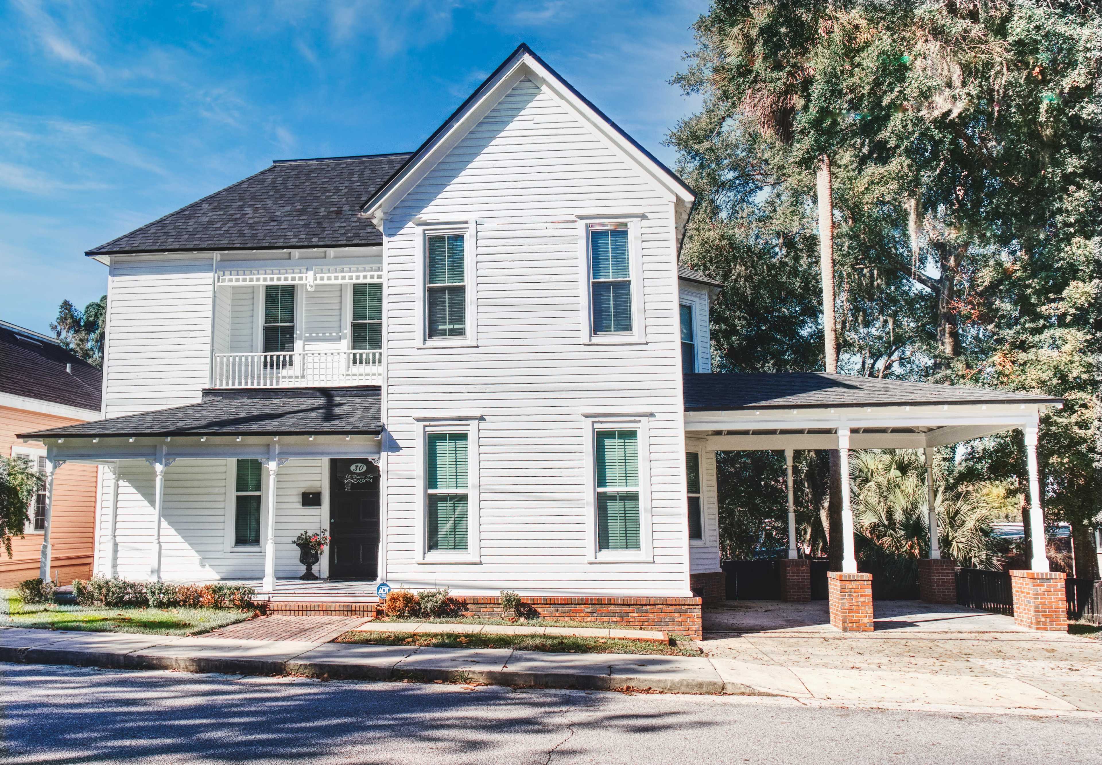 A two-story white wooden house with a black roof and a covered porch is situated on a street corner.