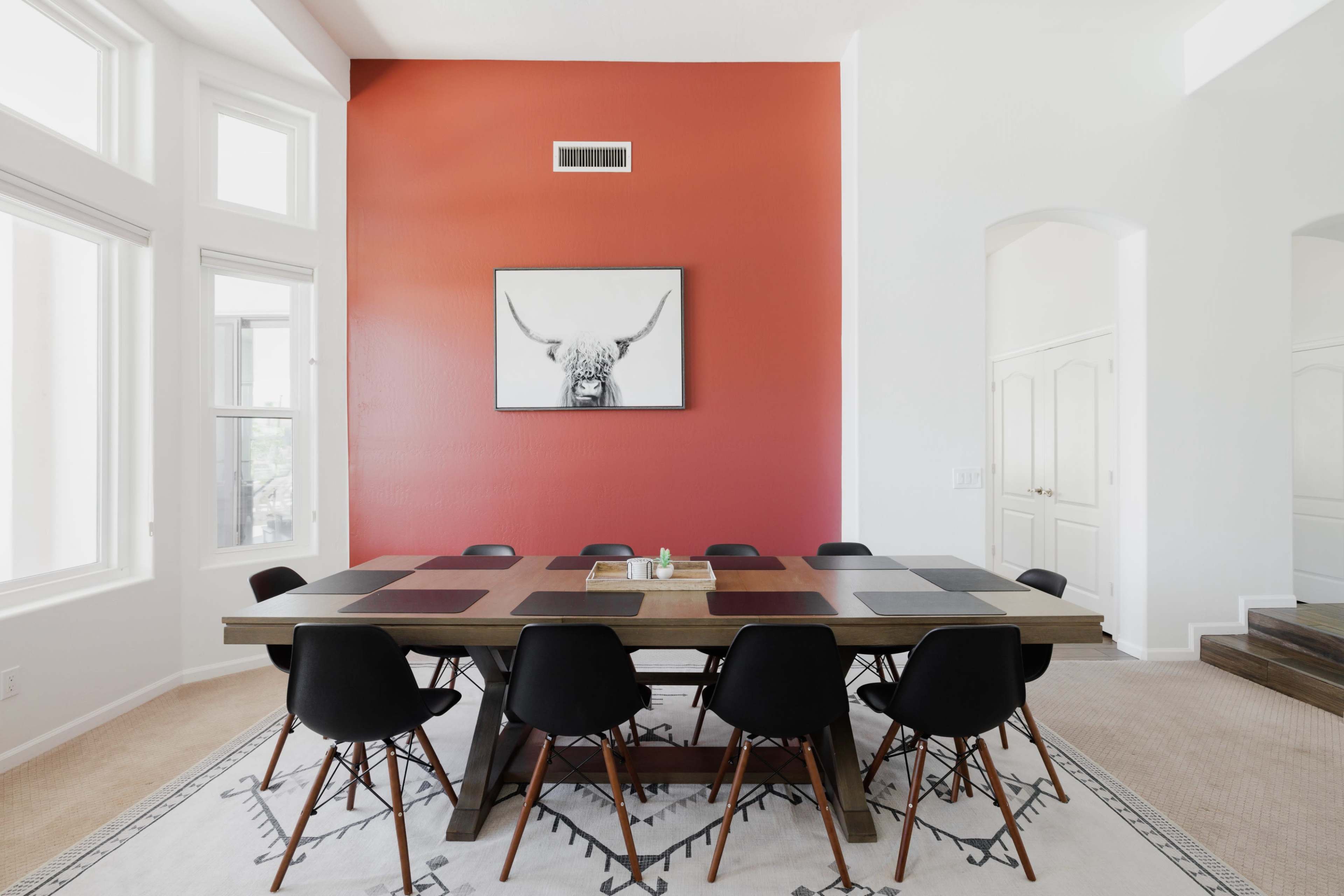 A long wooden dining table with black chairs is set against a red accent wall featuring a framed picture of a bull's head.