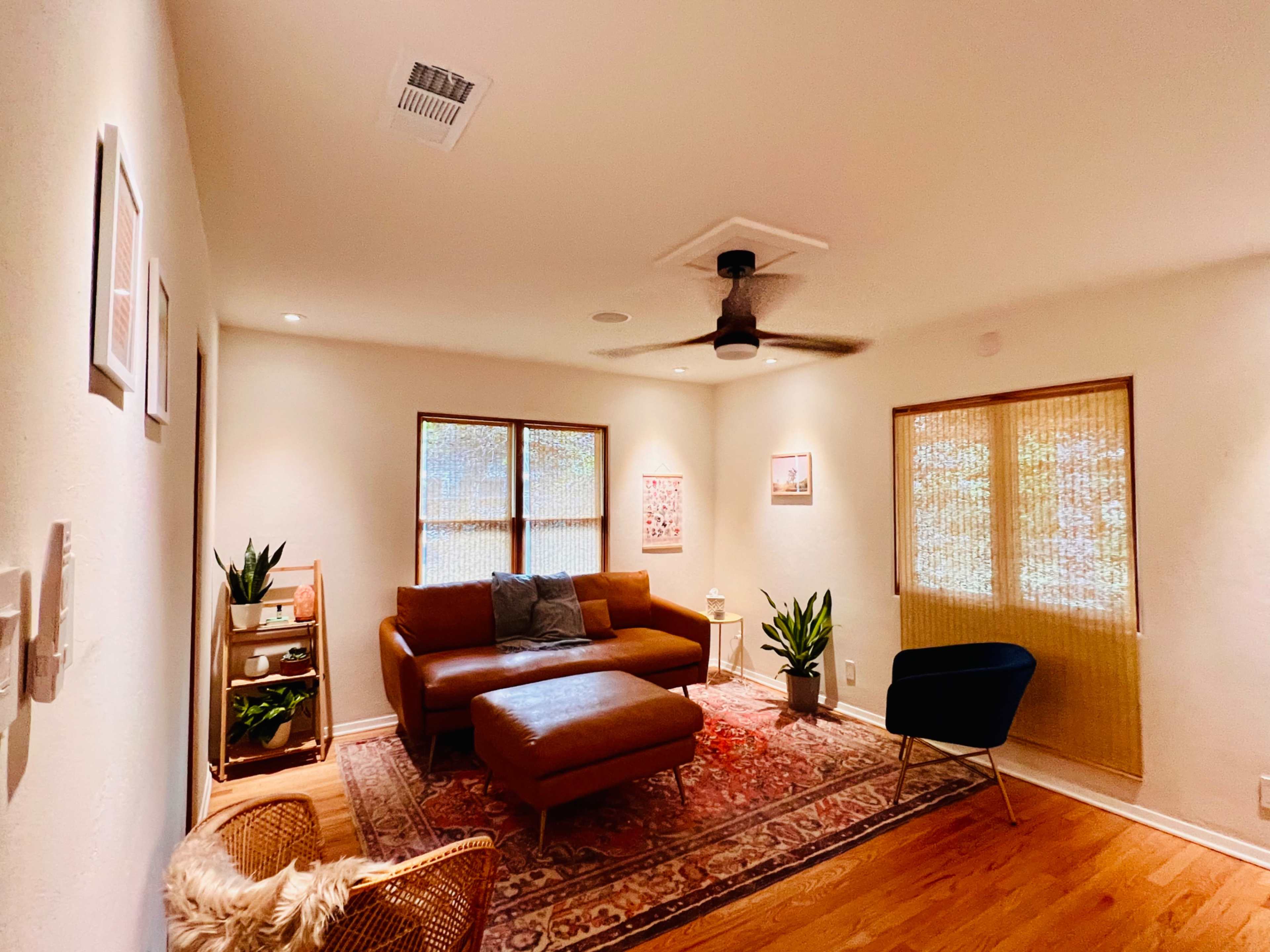 A cozy living room with a brown leather sofa, a coffee table, and two windows covered with bamboo shades, surrounded by indoor plants and a patterned rug.