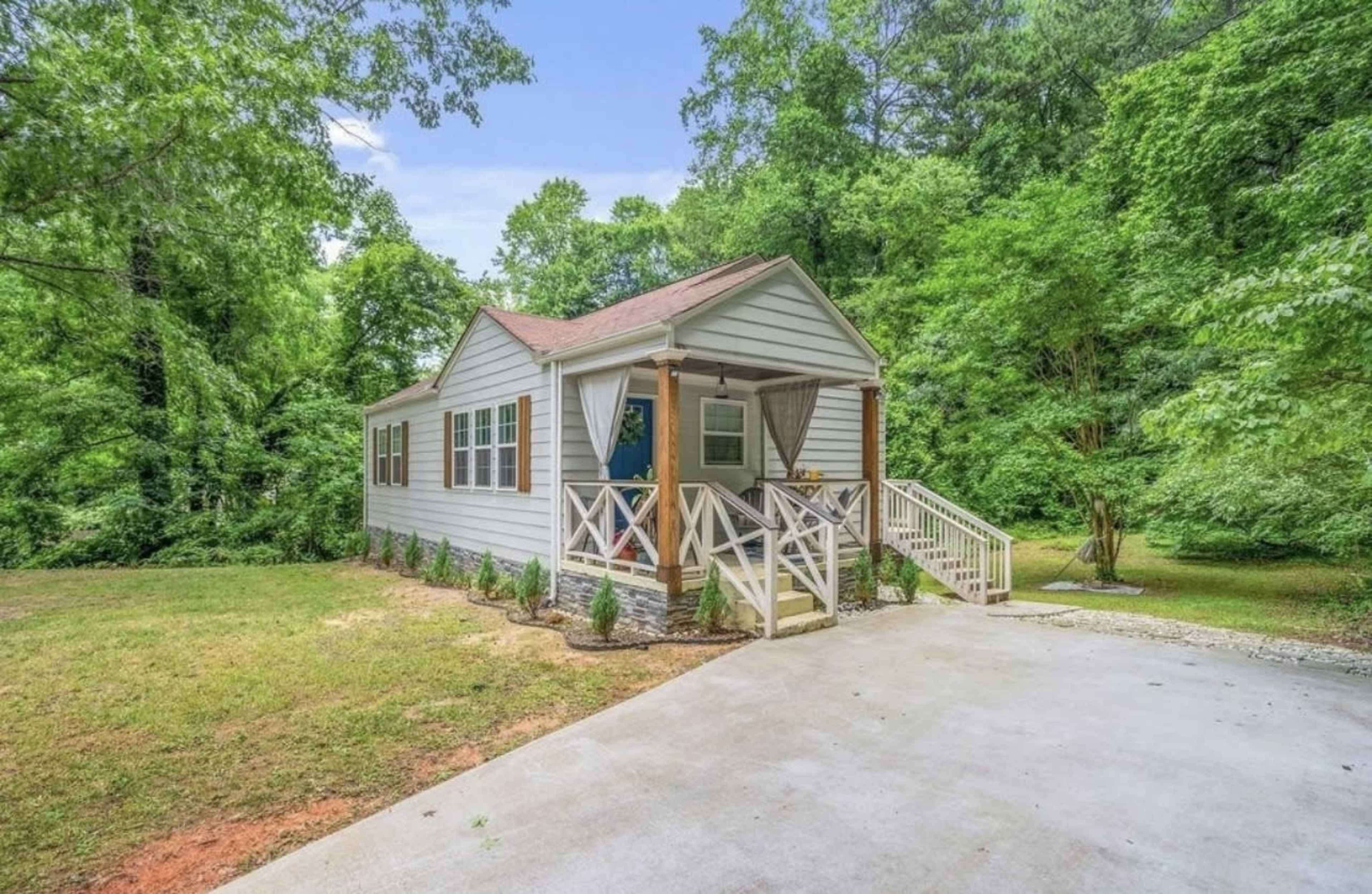 A single-story house with a front porch is situated on a landscaped lot surrounded by trees.