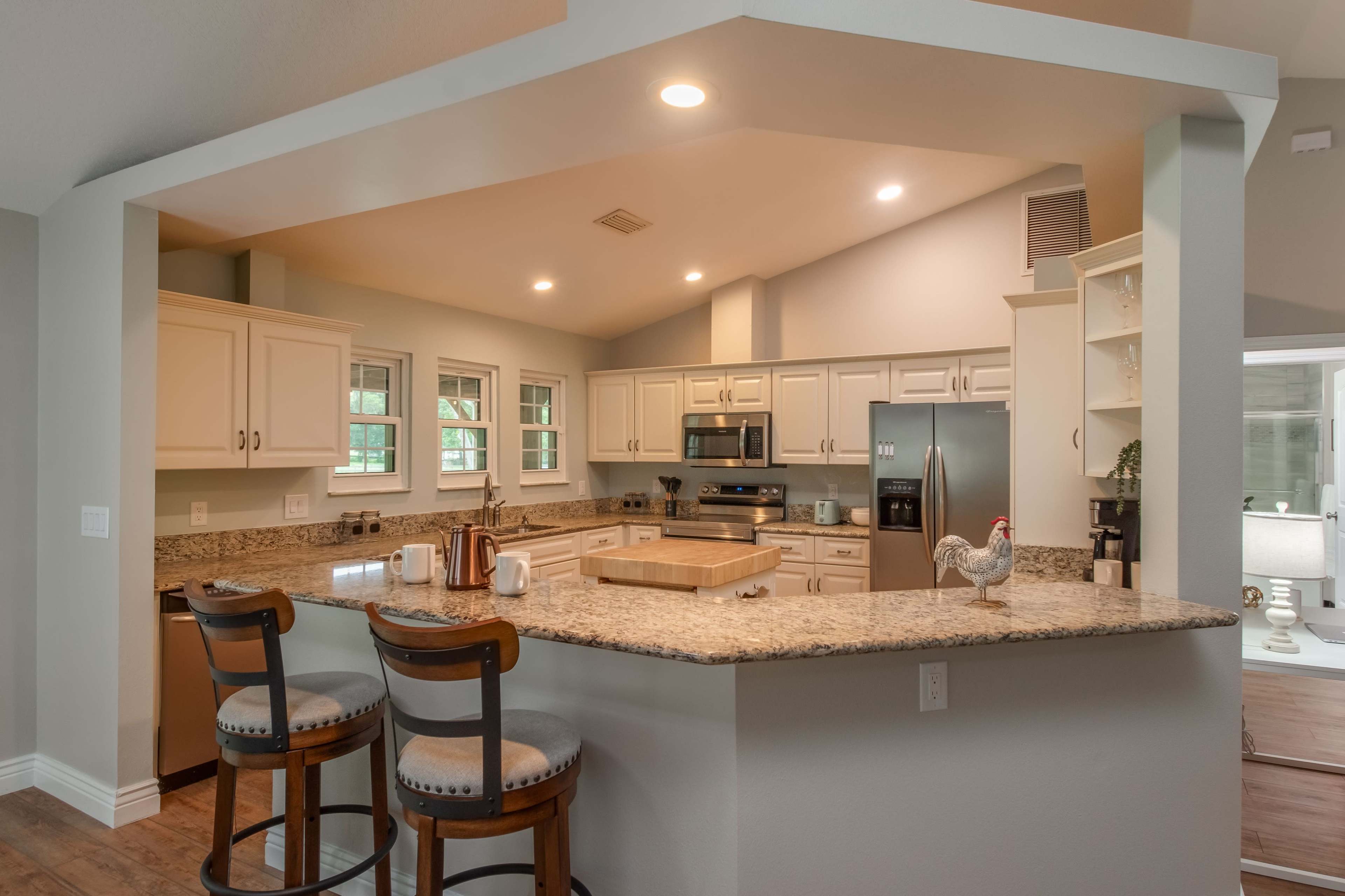 A modern kitchen with granite countertops, white cabinetry, and stainless steel appliances, featuring a breakfast bar with two stools.