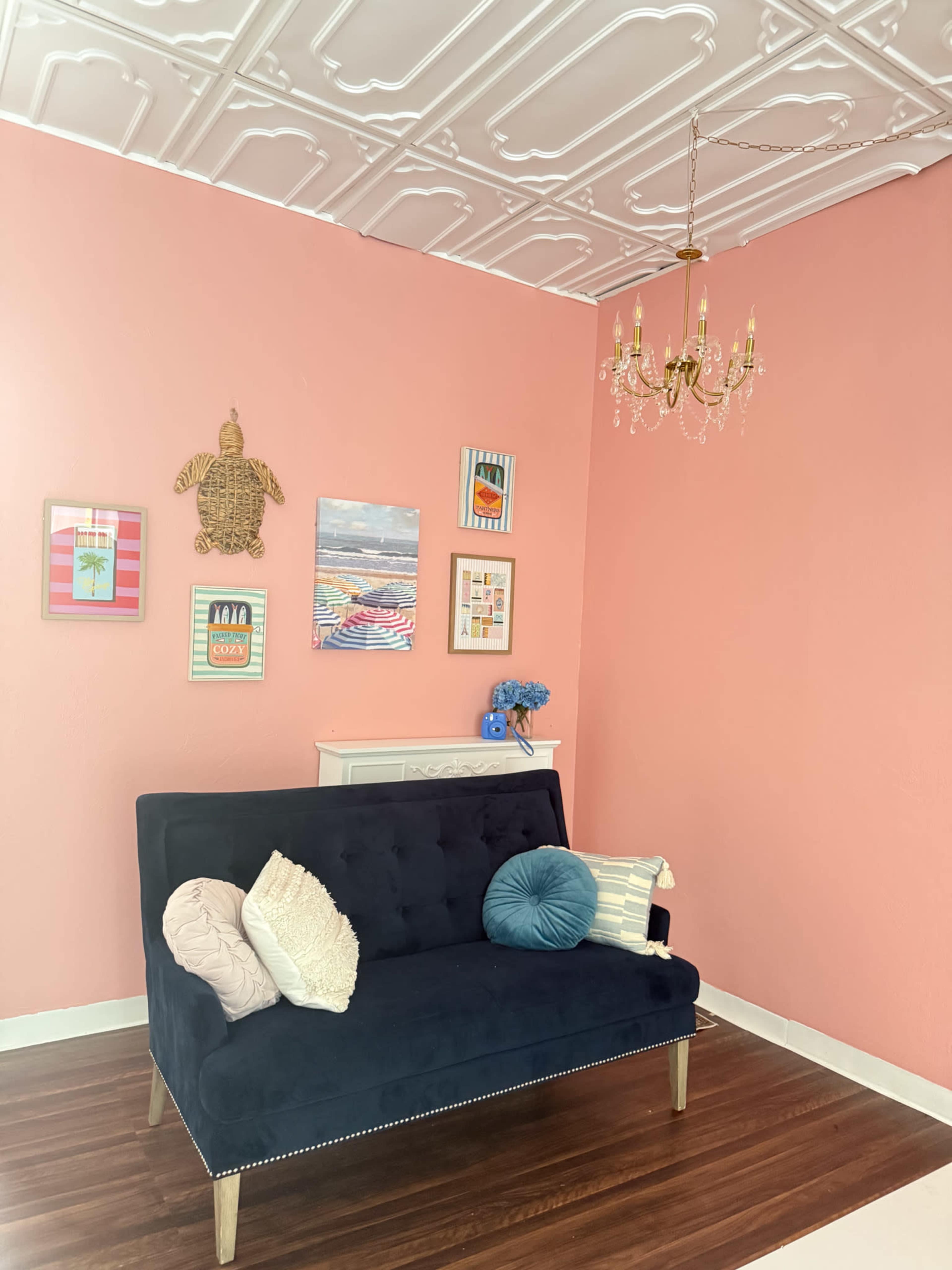 The image shows a cozy room with a pink wall, a navy blue sofa adorned with pillows, and several framed artworks hanging above a white cabinet.