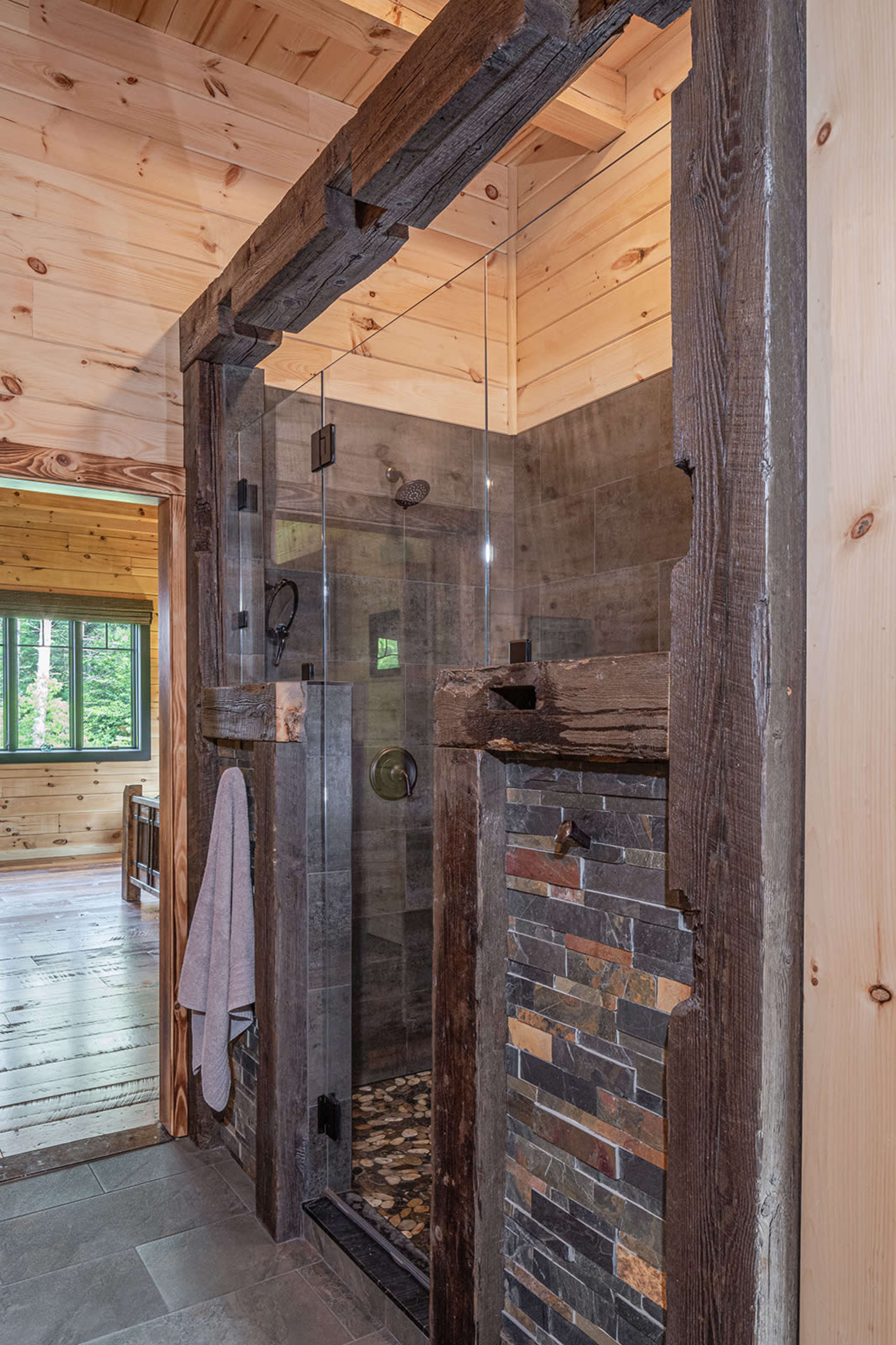 A modern shower enclosure with clear glass walls, framed by wooden beams, and featuring a stone accent wall and pebble floor.