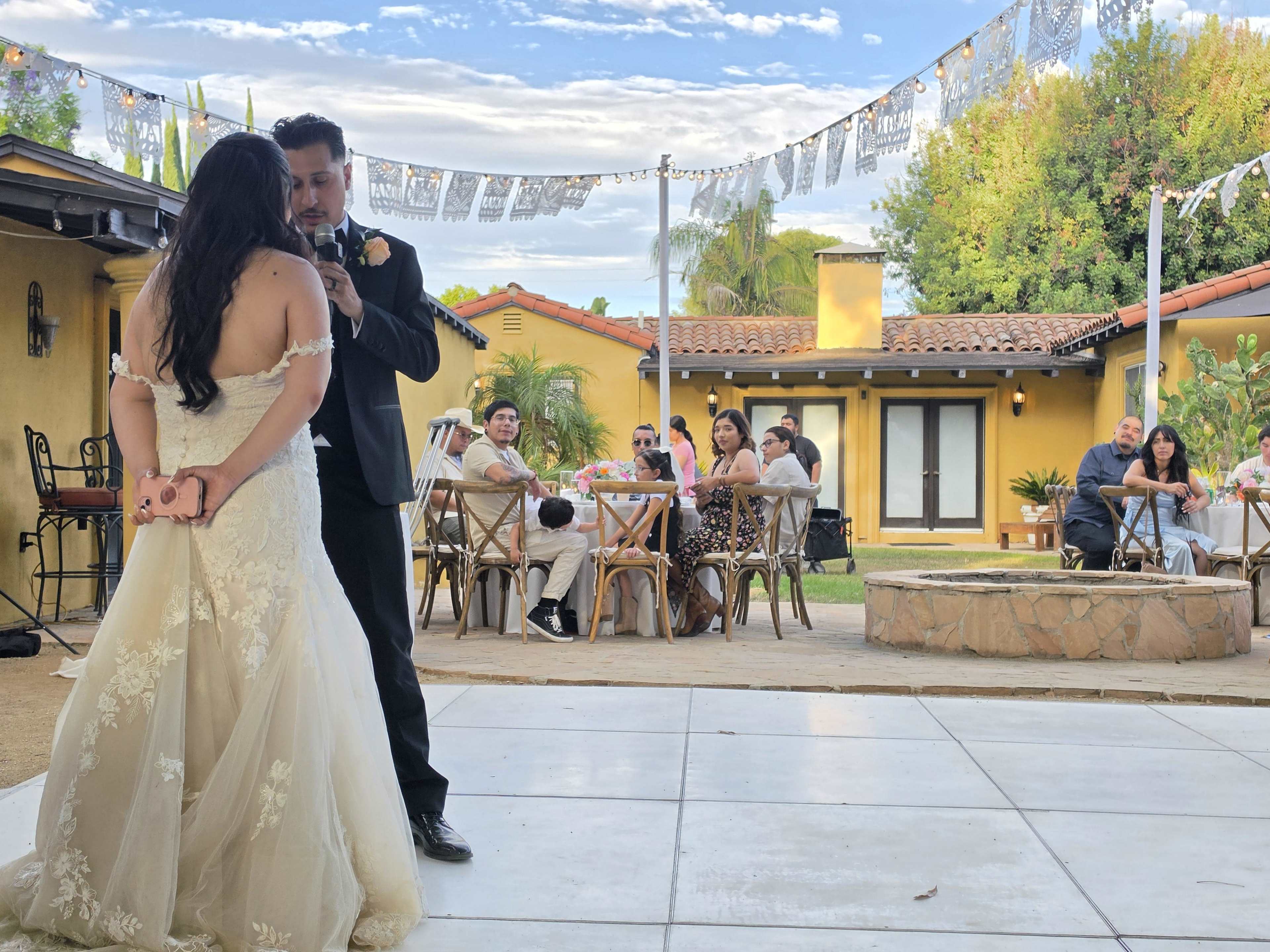 A couple stands facing each other during their wedding ceremony in a spacious outdoor venue, surrounded by guests seated at tables under decorative banners.