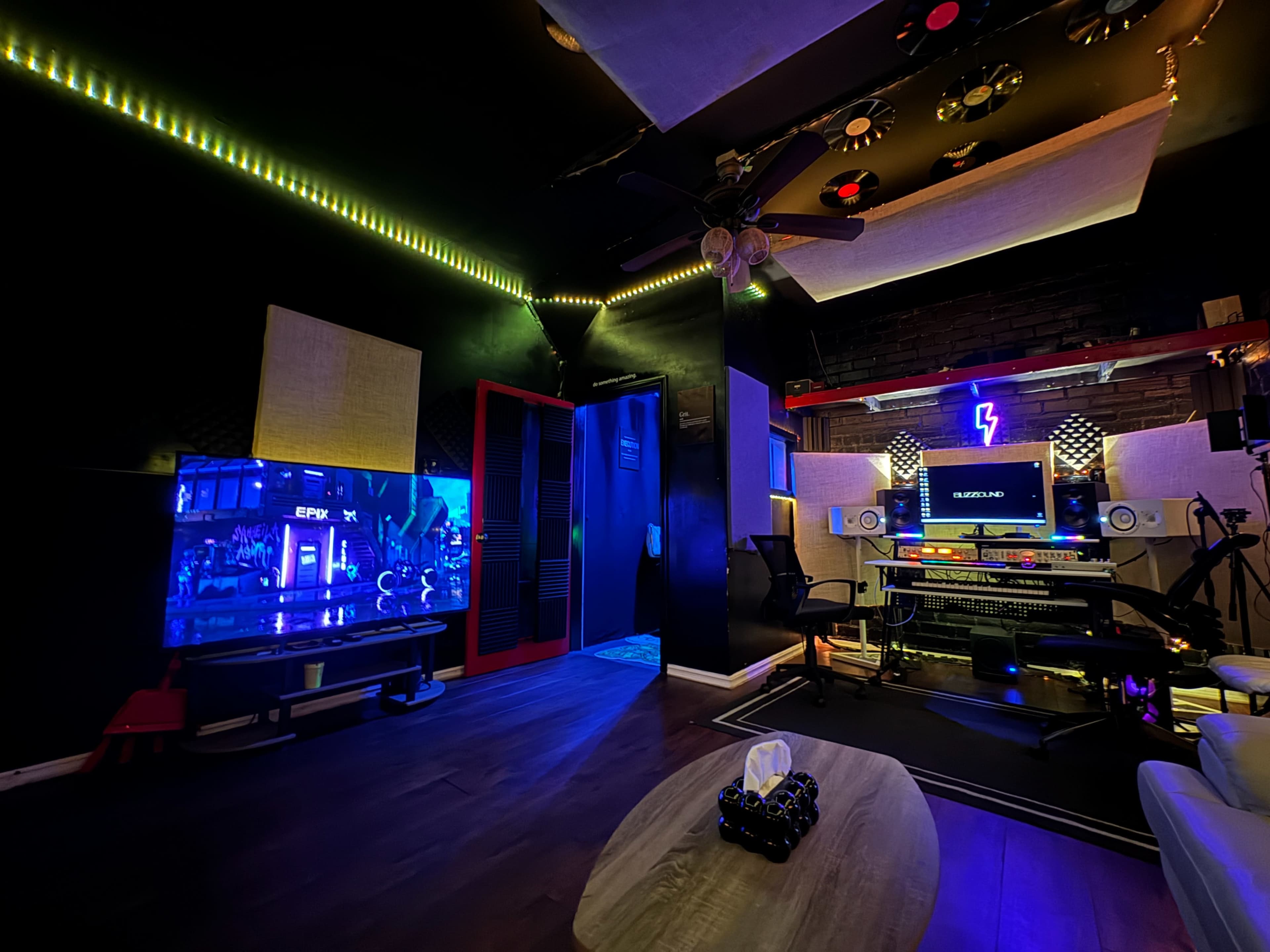 A dimly lit recording studio features a large screen, audio equipment, and mood lighting.