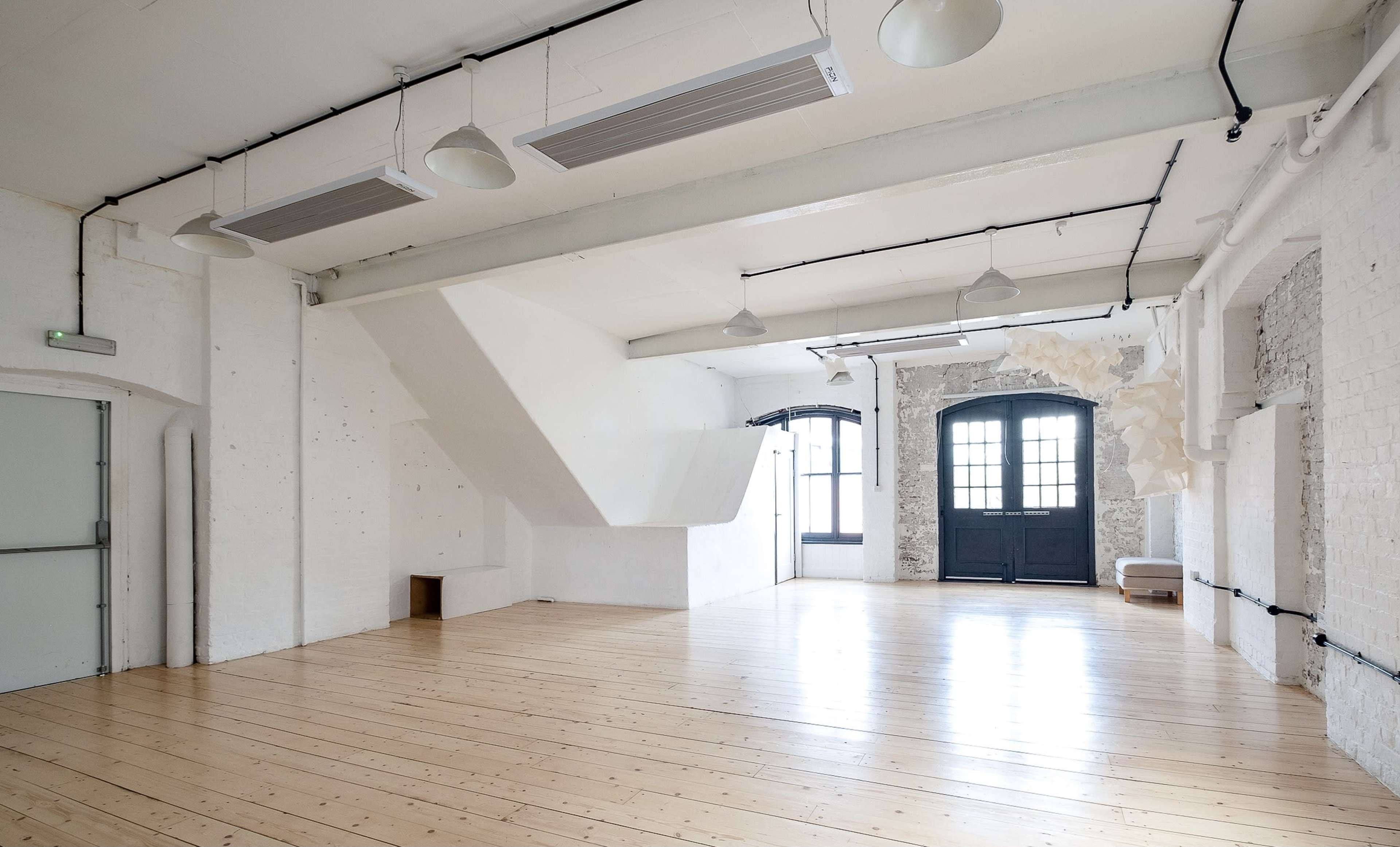 A bright, spacious room with white walls, wooden flooring, and large windows, featuring minimal furnishings and distinctive architectural elements.