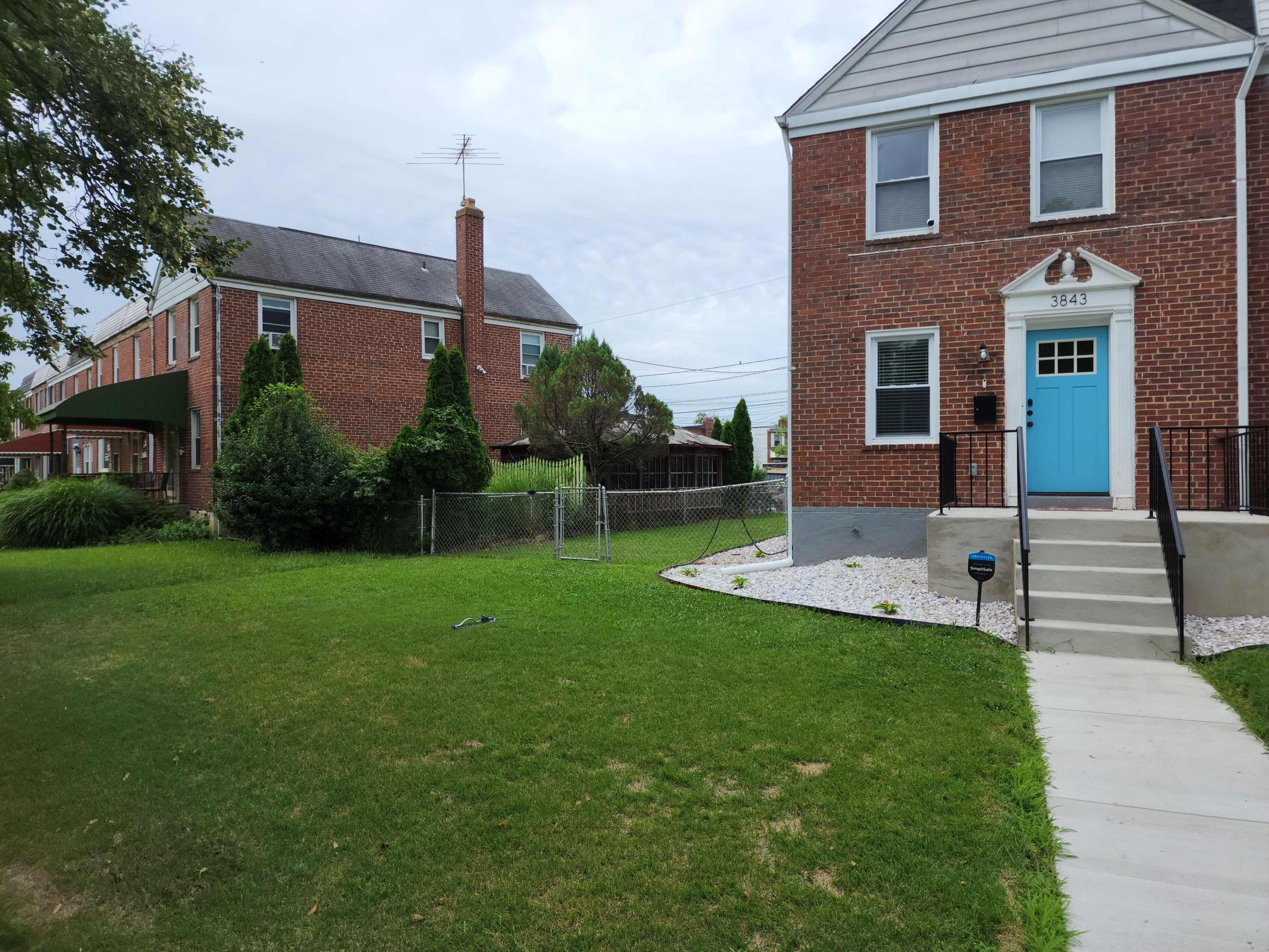 The image shows a brick house with a blue front door, a concrete walkway, and a well-maintained lawn, alongside another red-brick building in the background.