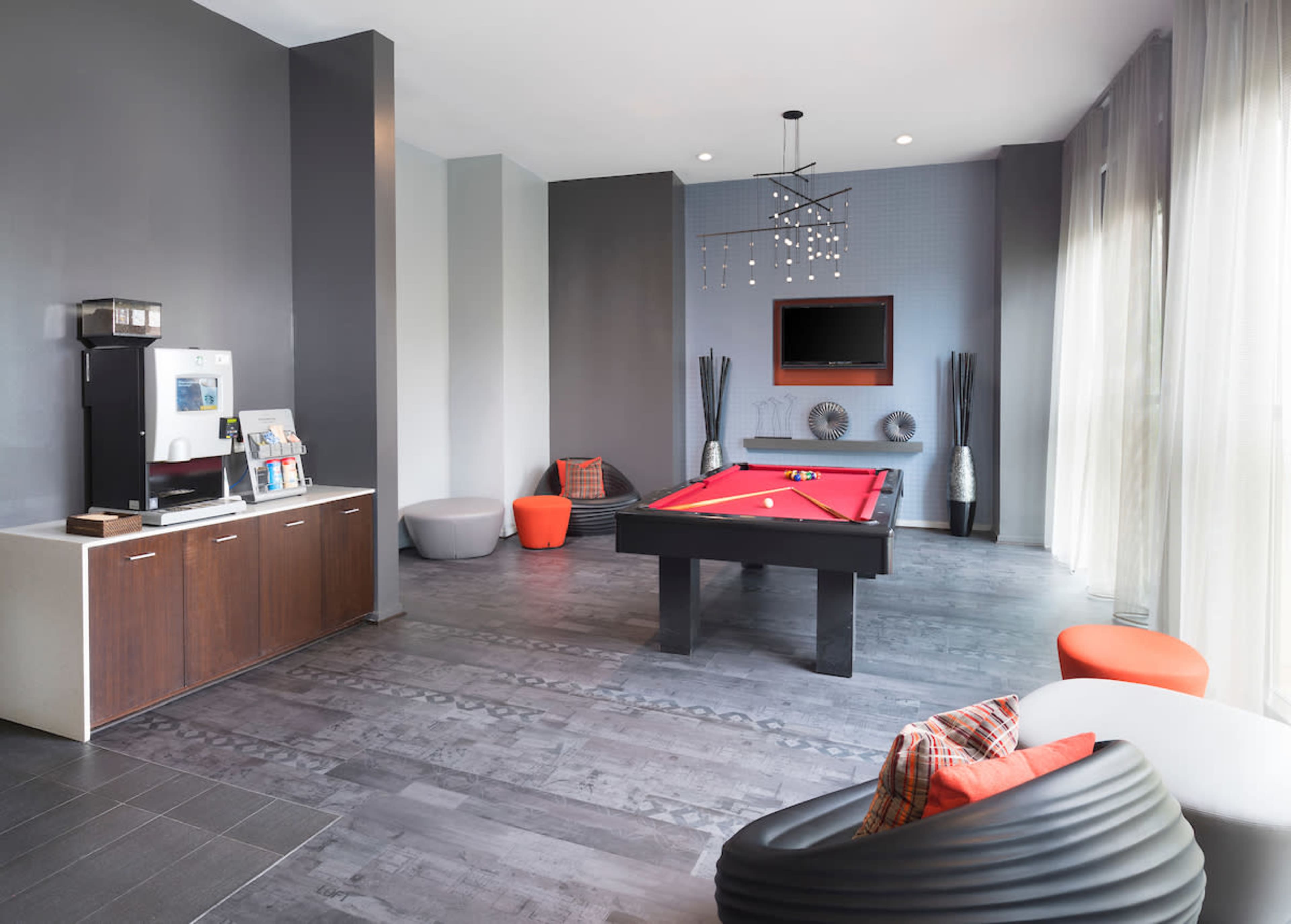The image shows a modern game room featuring a red pool table, a television on the wall, and a seating area with contemporary furniture.