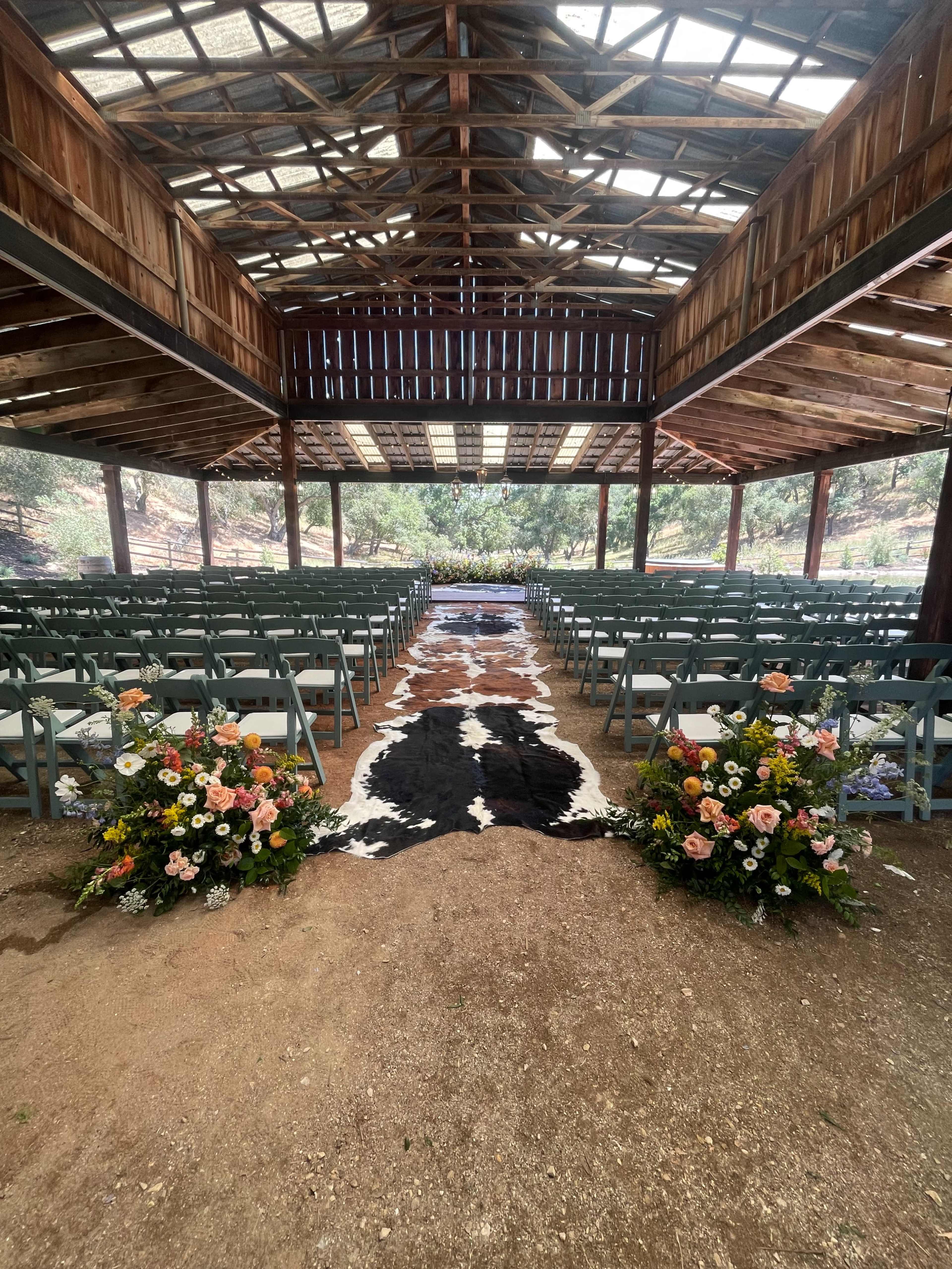 A rustic outdoor venue features rows of chairs facing a central aisle adorned with a cowhide rug and floral arrangements on either side.