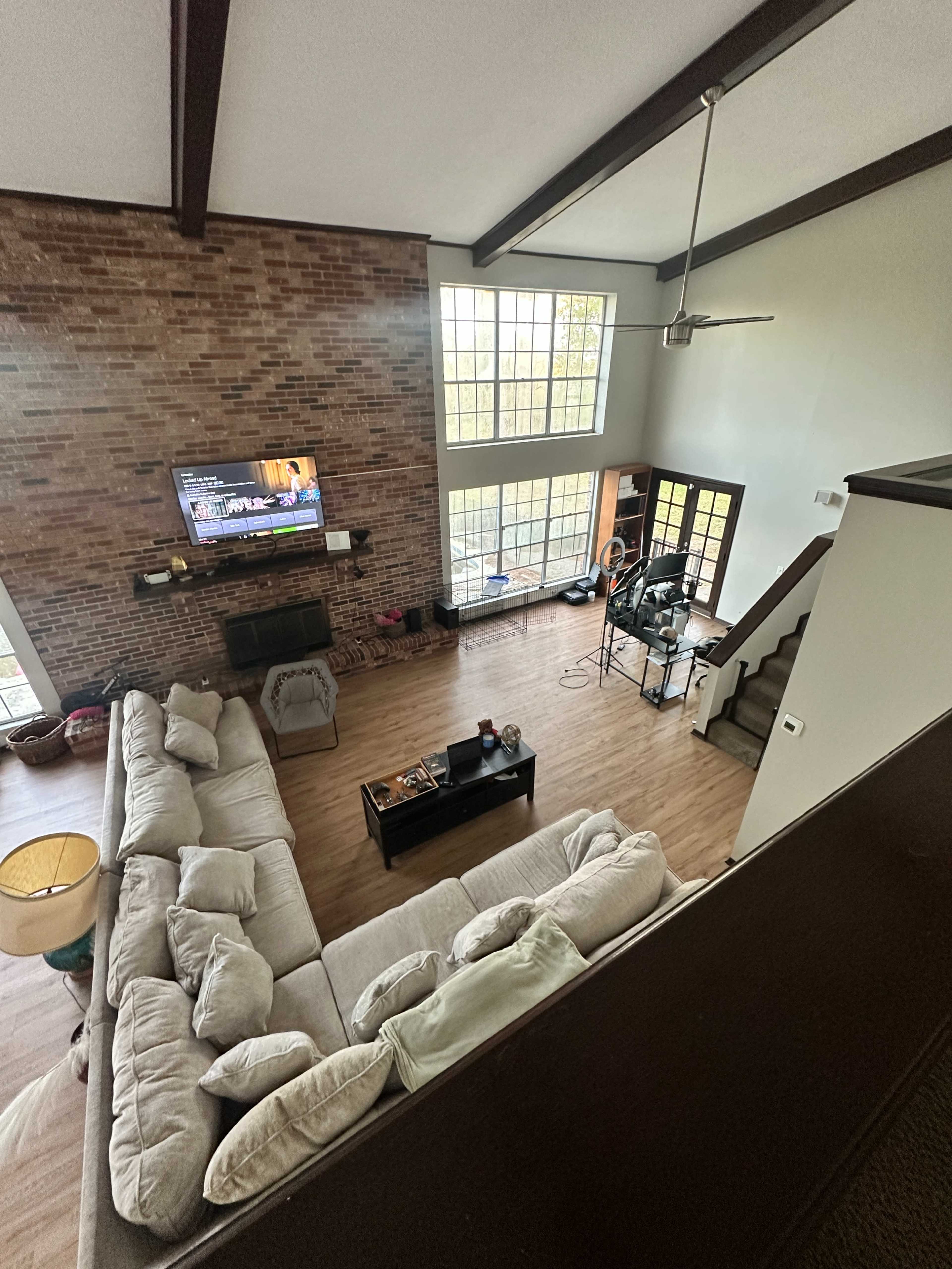 The image shows a spacious living room with a large sectional sofa, a television mounted on a brick wall, and large windows allowing natural light to enter.