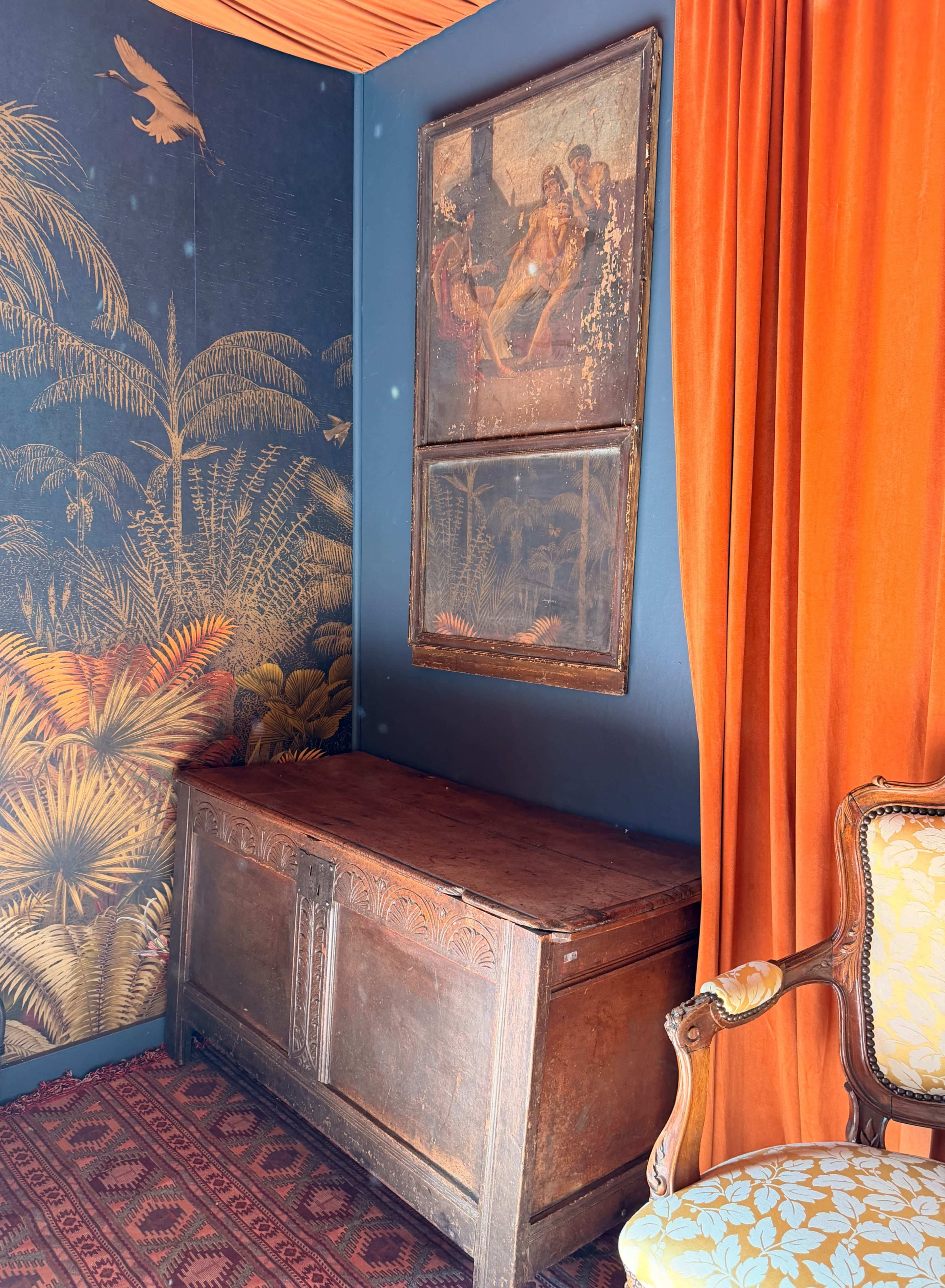 The image shows a room with a wooden chest, a framed painting on the wall, and orange curtains draping beside a patterned chair.
