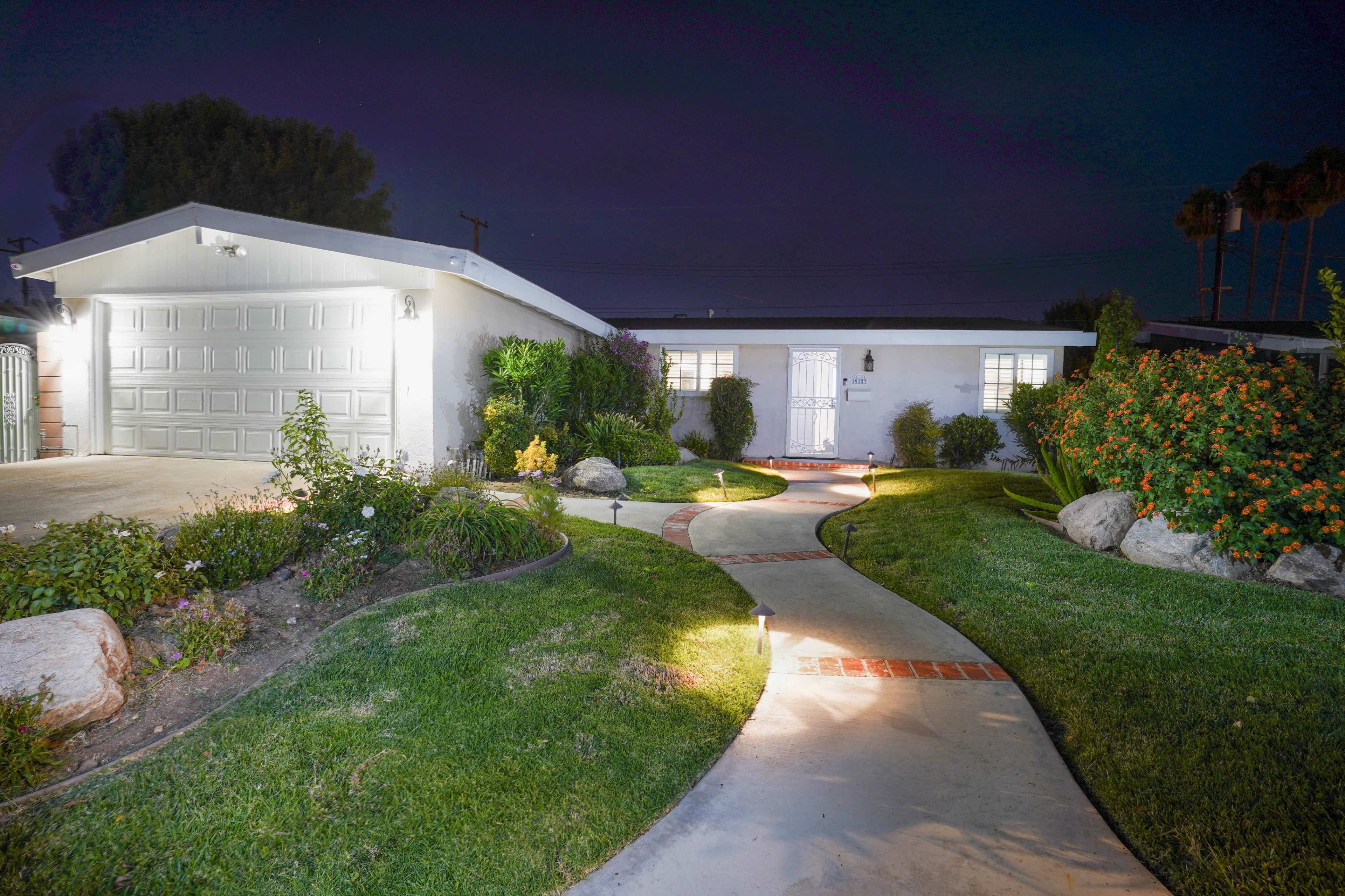 A well-lit pathway leads to a single-story home with a garage, surrounded by manicured lawns and flower beds at night.