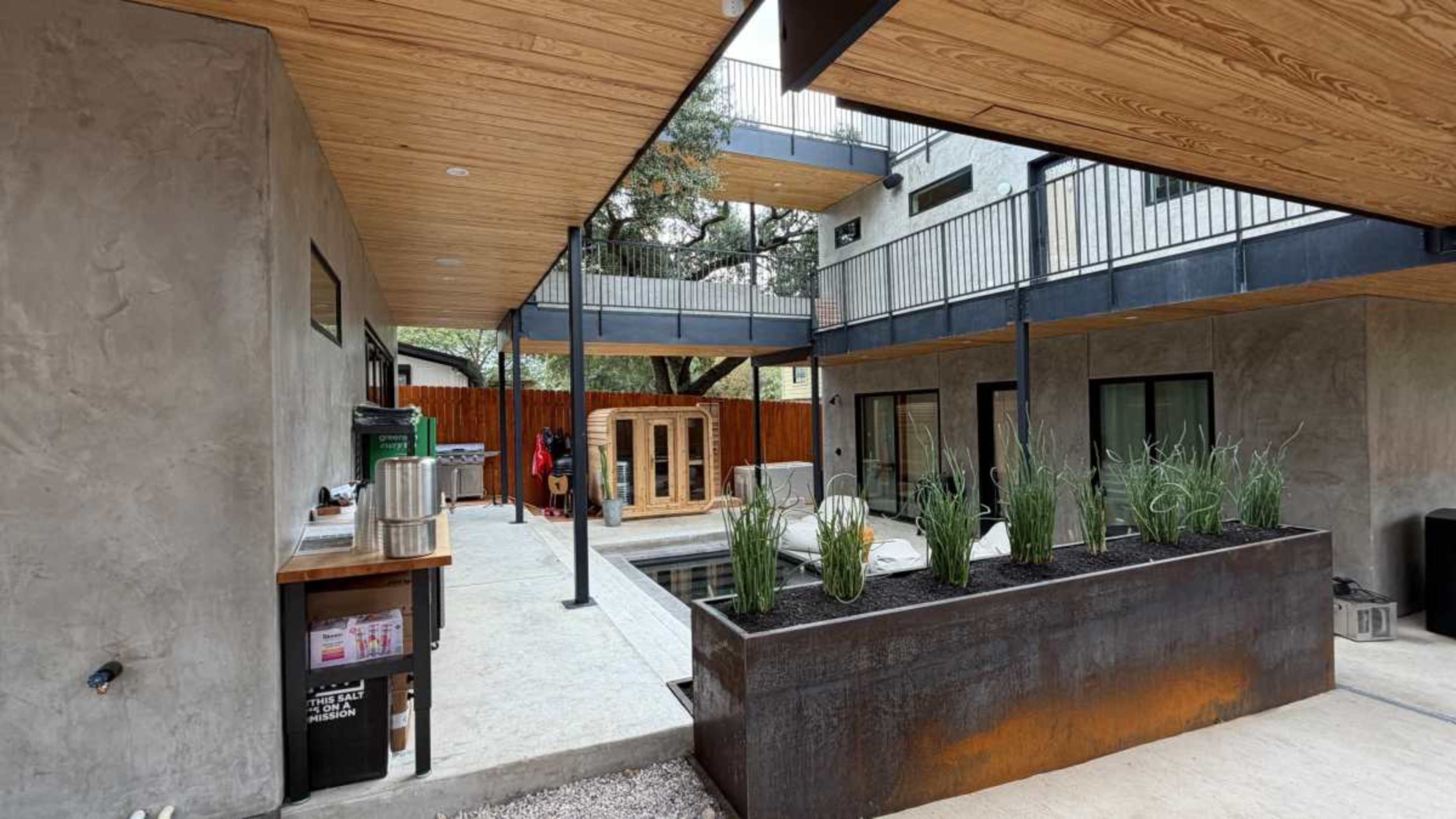 The image shows a modern outdoor space featuring a minimalist design, with a concrete patio, glass railings, and a sauna surrounded by raised planter beds.