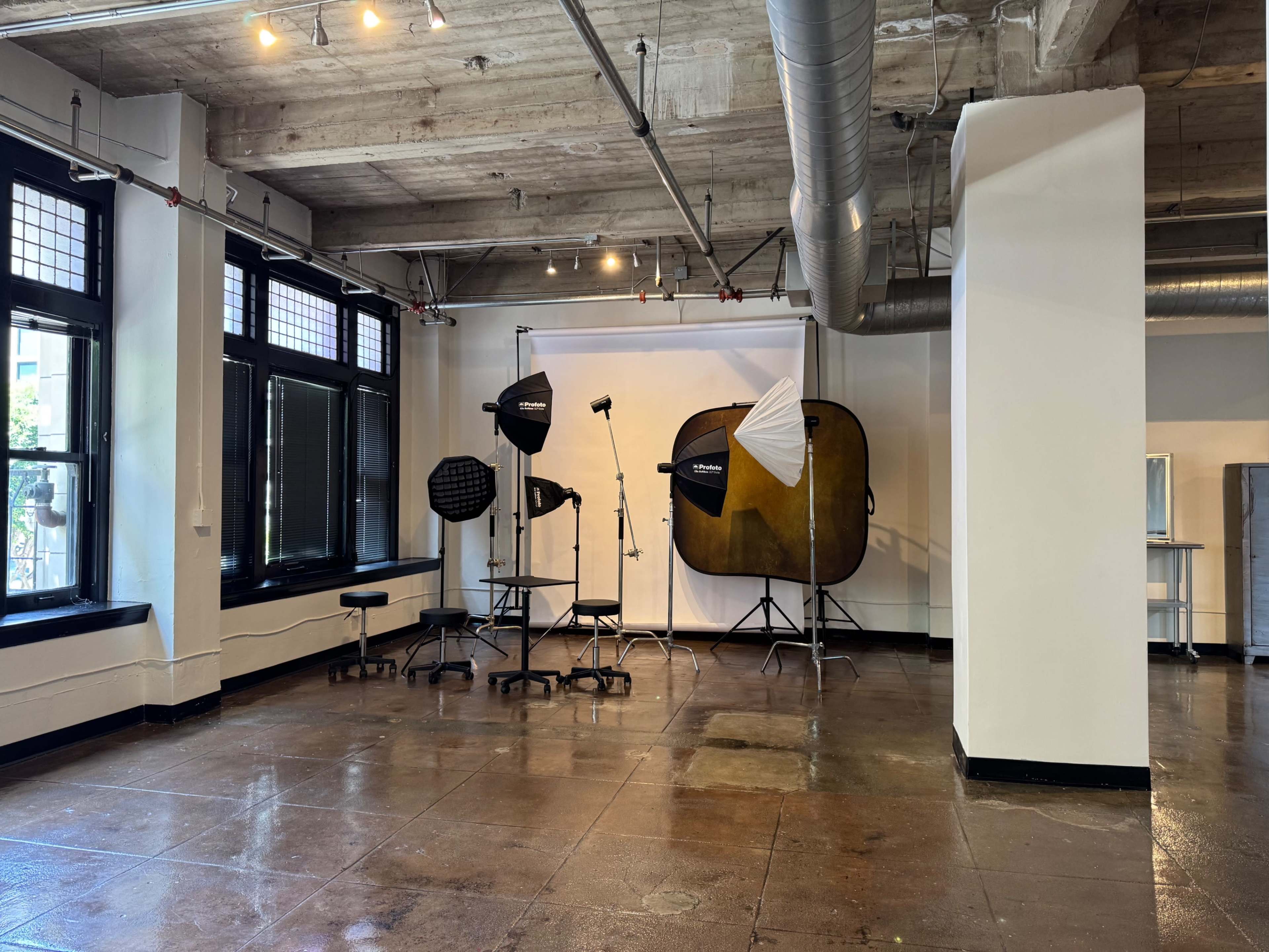The image shows a photography studio equipped with lighting gear, softboxes, and a backdrop set up in a spacious room with large windows.