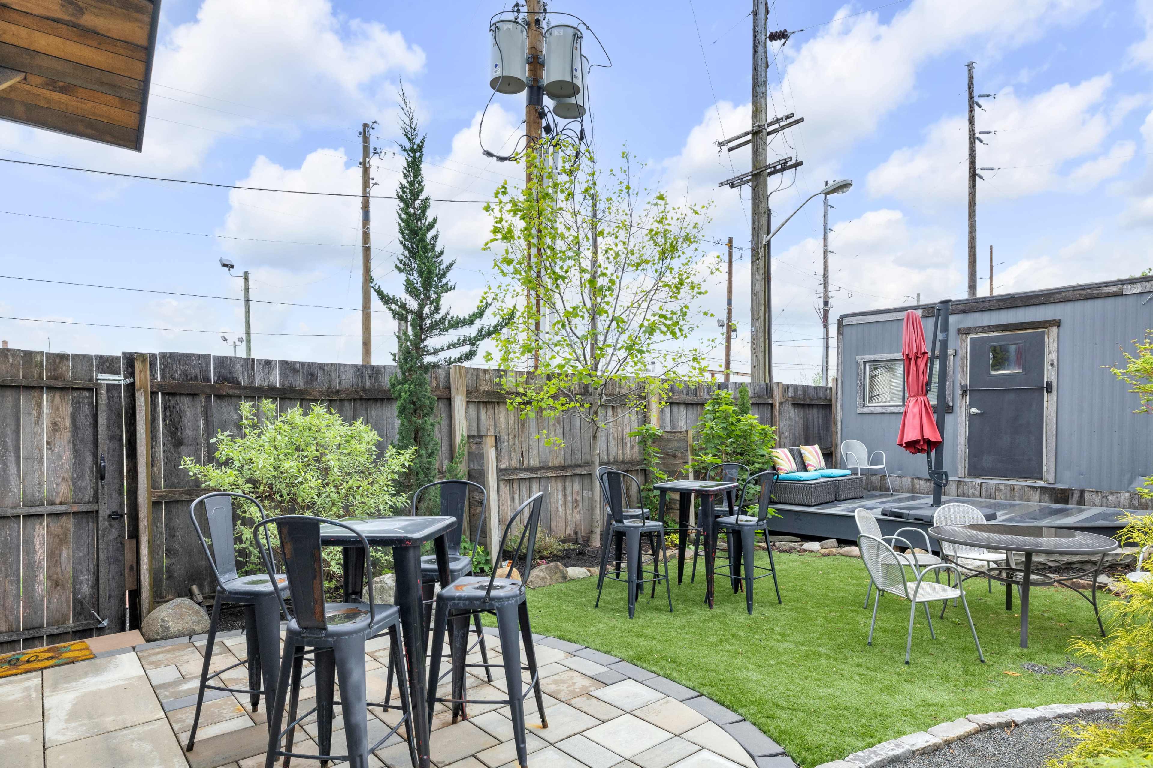 The image shows a landscaped outdoor patio area with several metal tables and chairs, surrounded by greenery and a wooden fence.