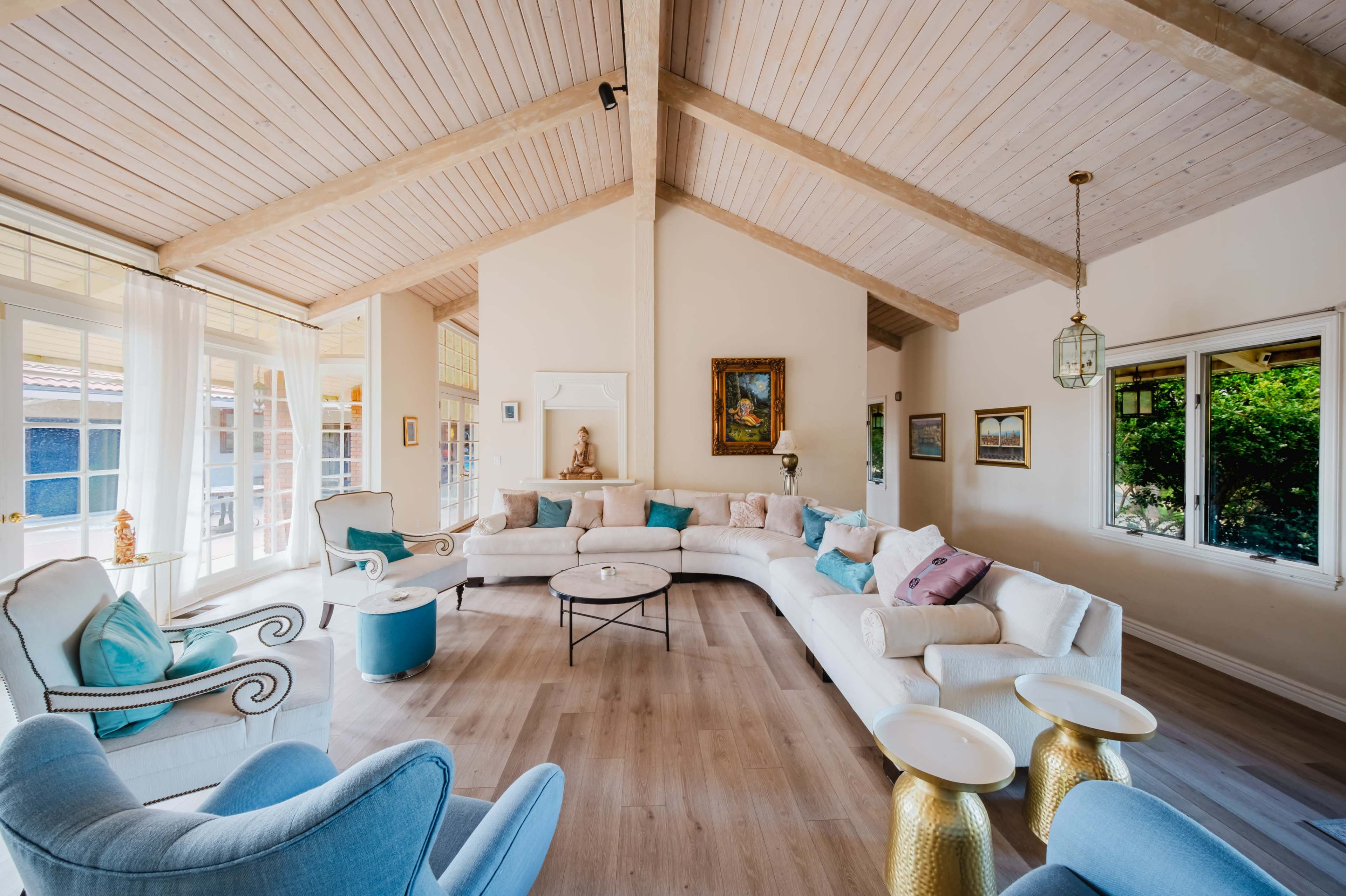 A spacious living room features a high wooden ceiling, light-colored walls, and a combination of white sofas accented with blue cushions, paired with gold side tables.