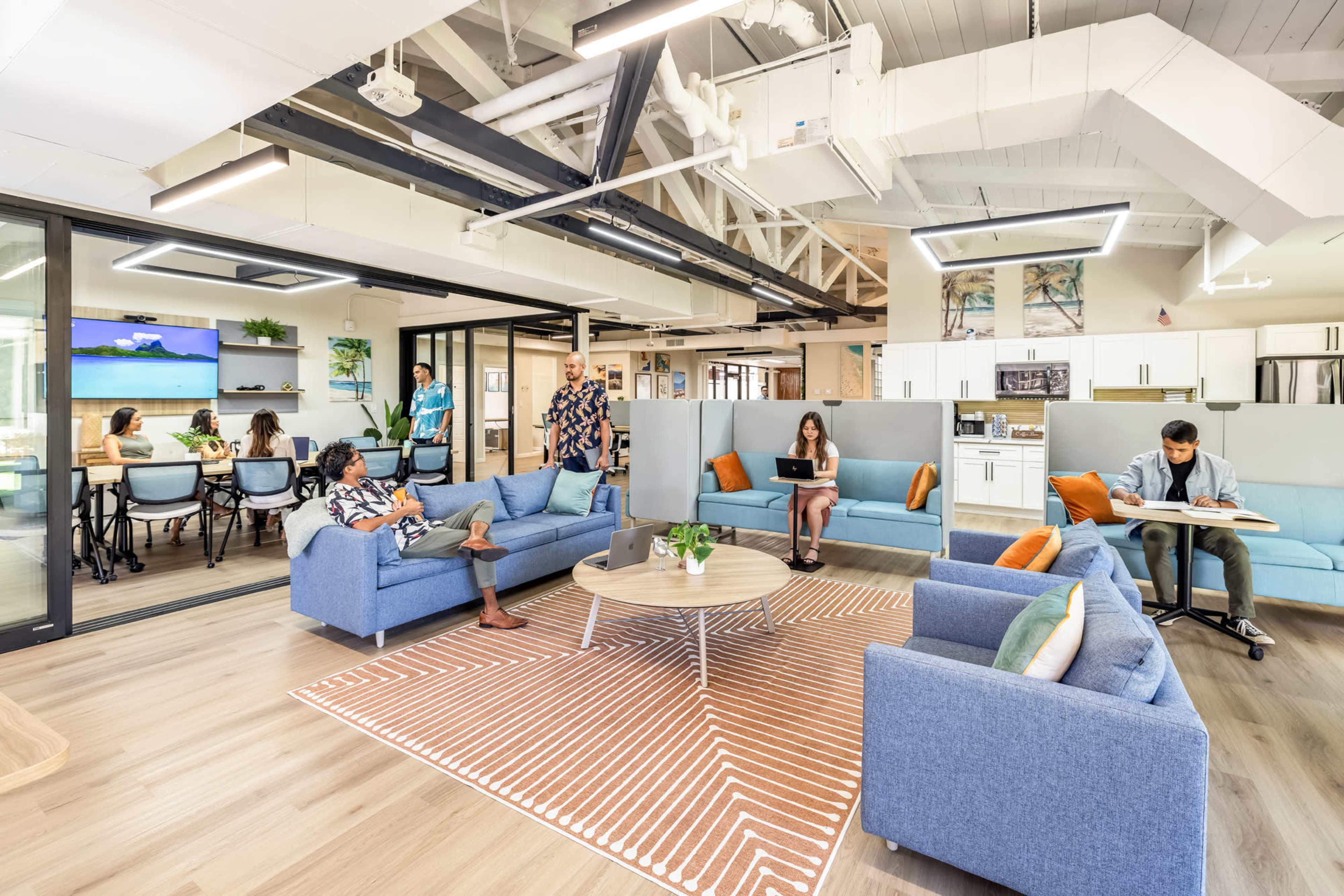A modern office space featuring blue couches, a central coffee table, and a mix of individuals working and collaborating in a bright environment.