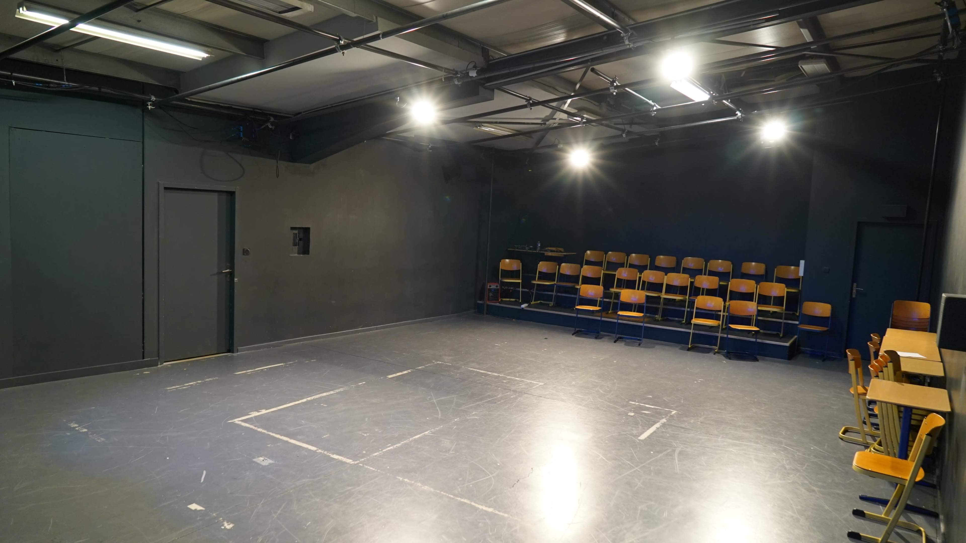 A sparsely furnished theater stage with a dark backdrop, illuminated by overhead lights and featuring rows of wooden chairs against one wall.