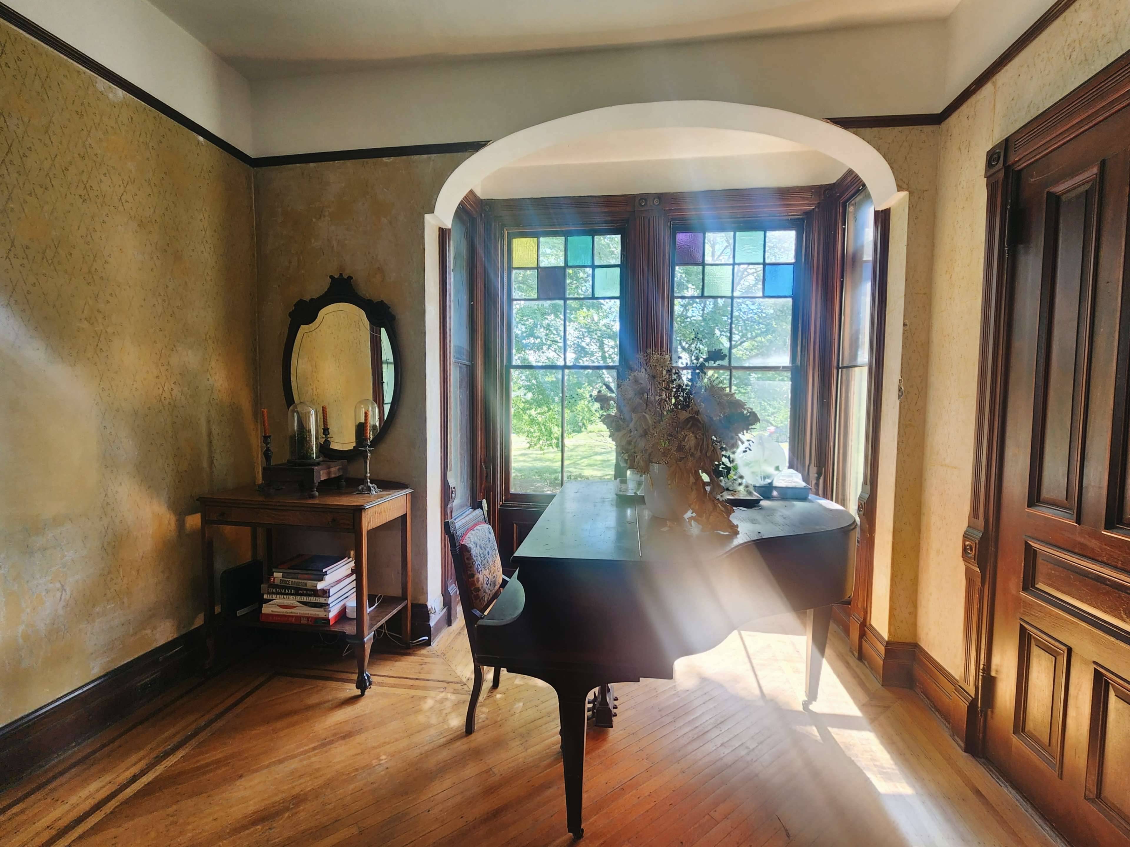 A grand piano sits in a bright room with large windows and vintage furnishings.