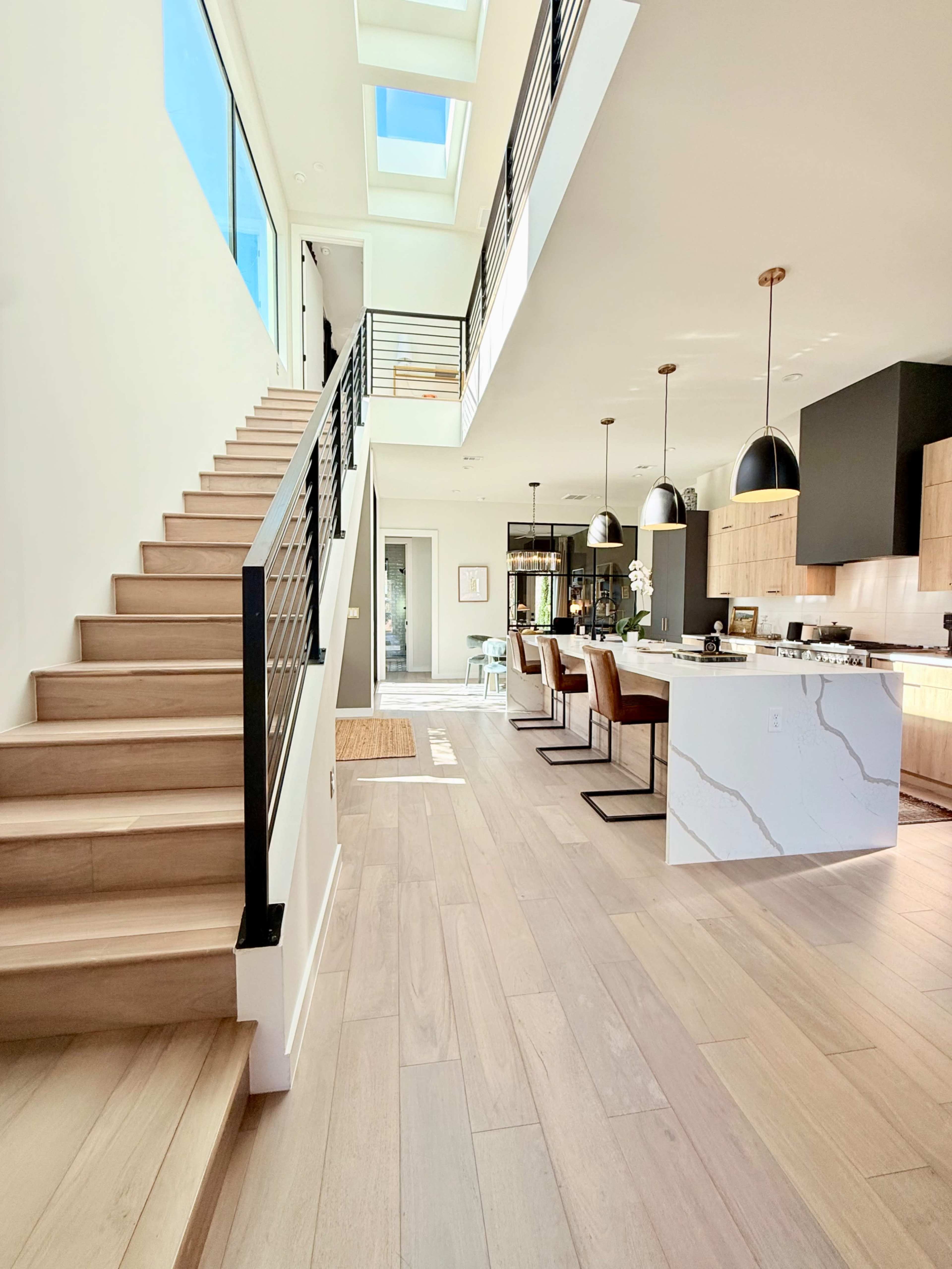 The image features a modern interior with a staircase leading to a second floor, an open kitchen and dining area, and large windows allowing natural light to fill the space.