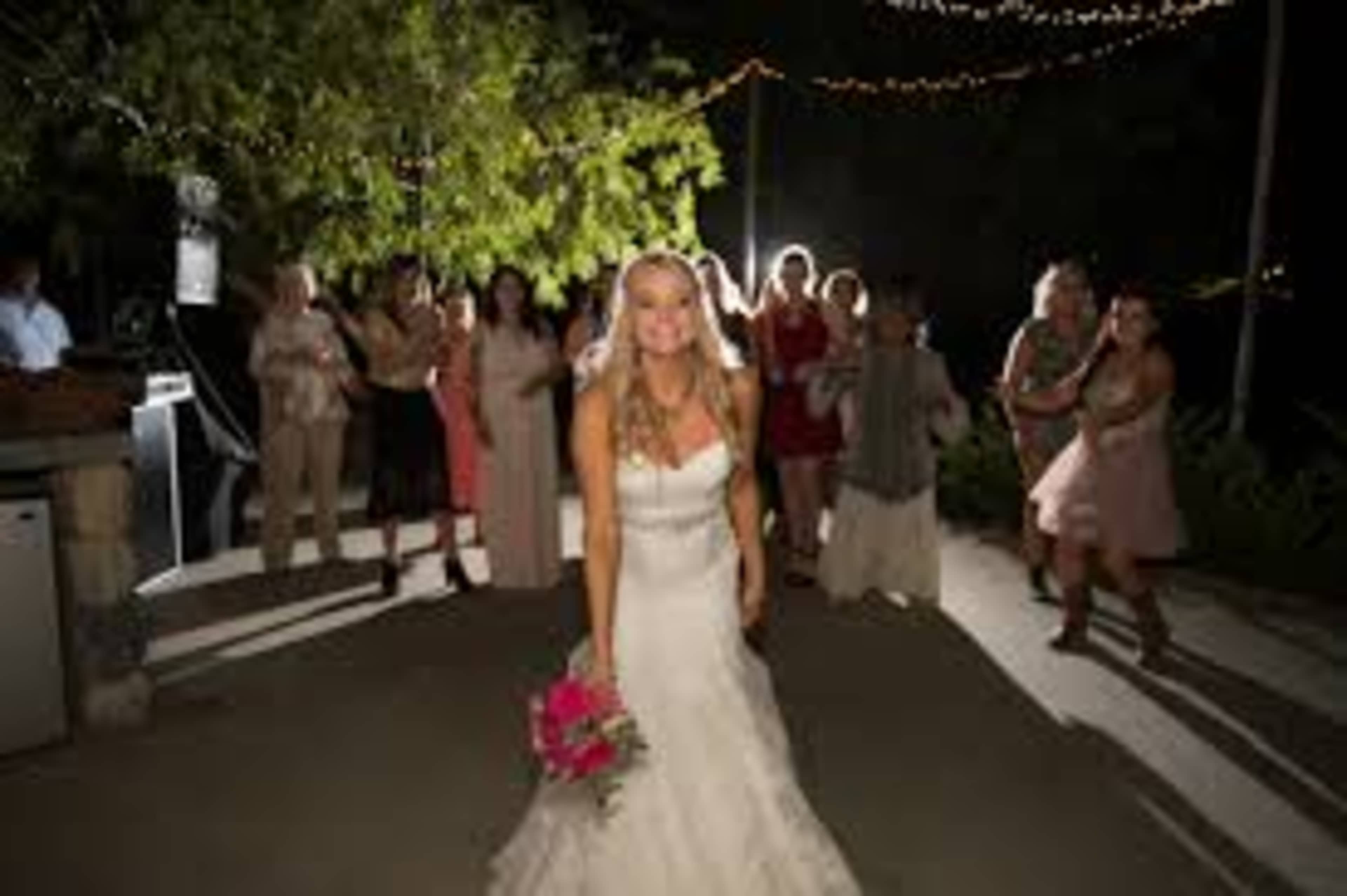A bride in a white wedding dress stands in front of a group of women holding a bouquet, with string lights overhead creating a festive atmosphere.