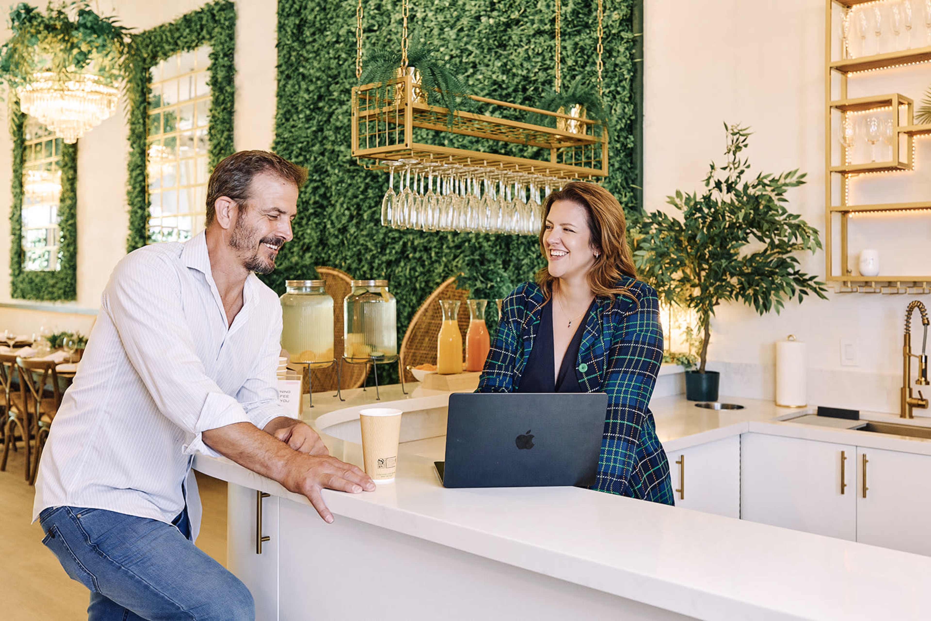 A man and a woman are chatting at a bright, modern café with greenery on the walls and drinks displayed behind them.