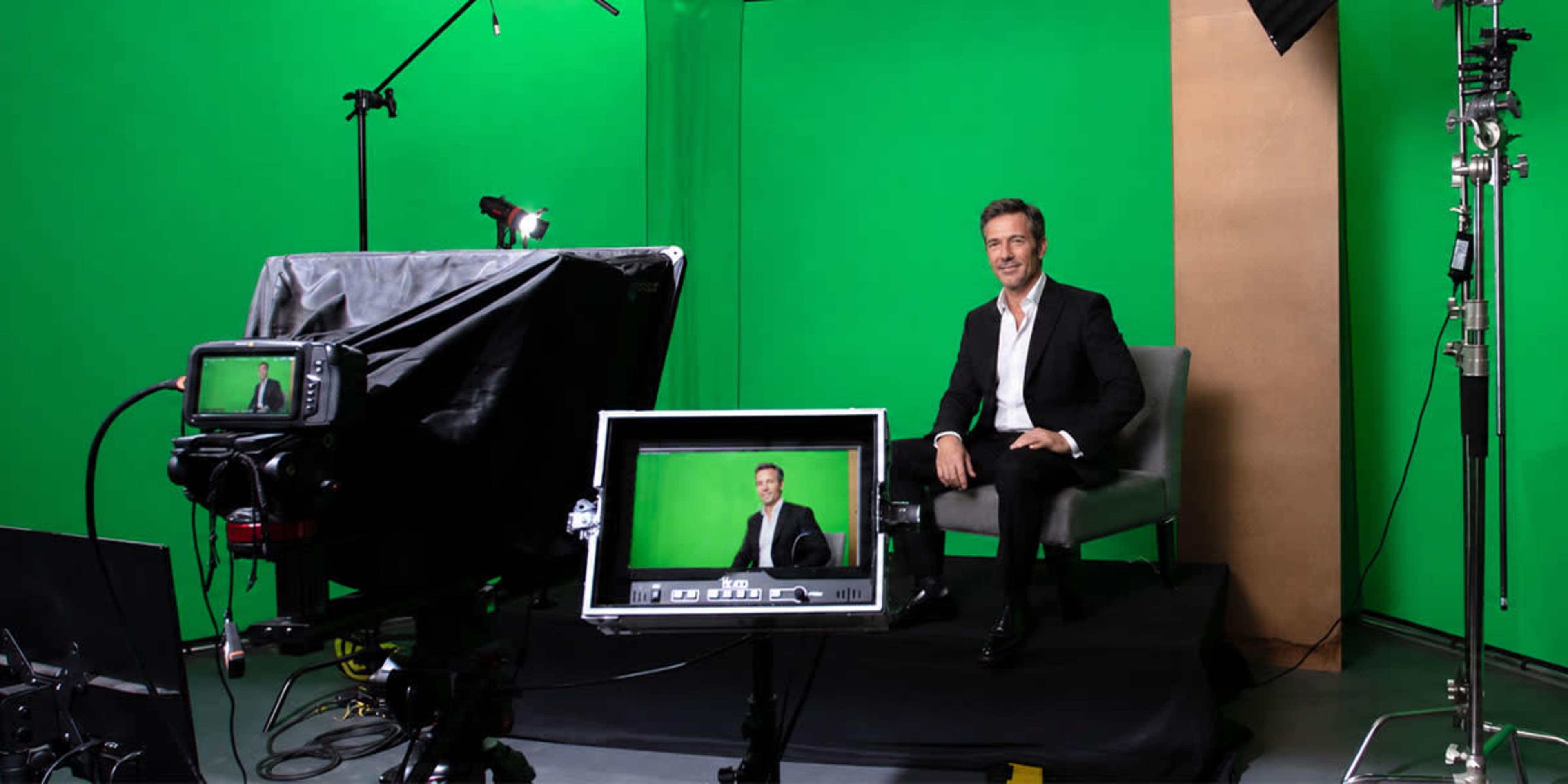 A man in a suit is seated on a chair in front of a green backdrop, while cameras and lighting equipment are positioned around him.