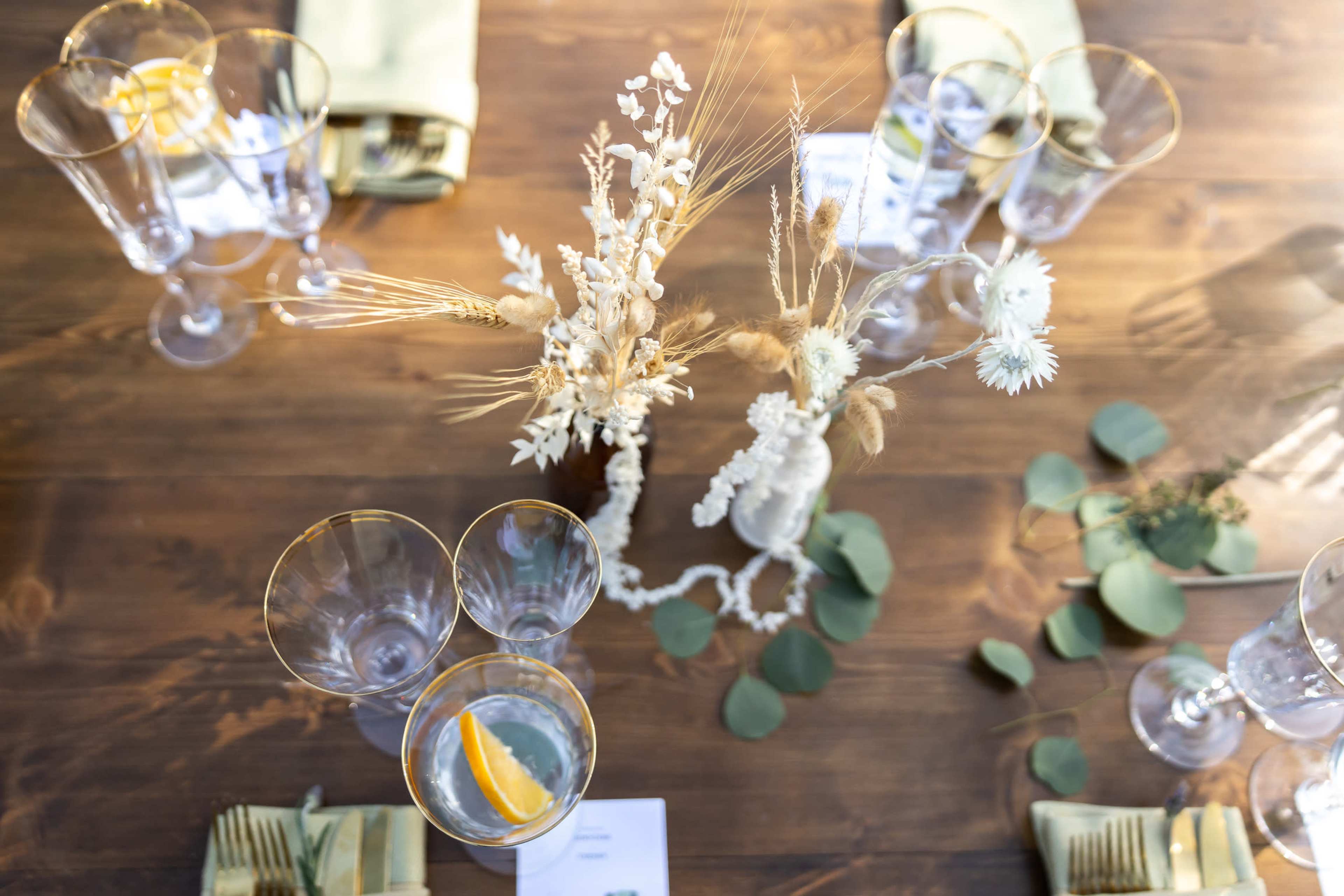 The image features a decorative table setting with glassware, floral arrangements, and greenery arranged on a wooden surface.