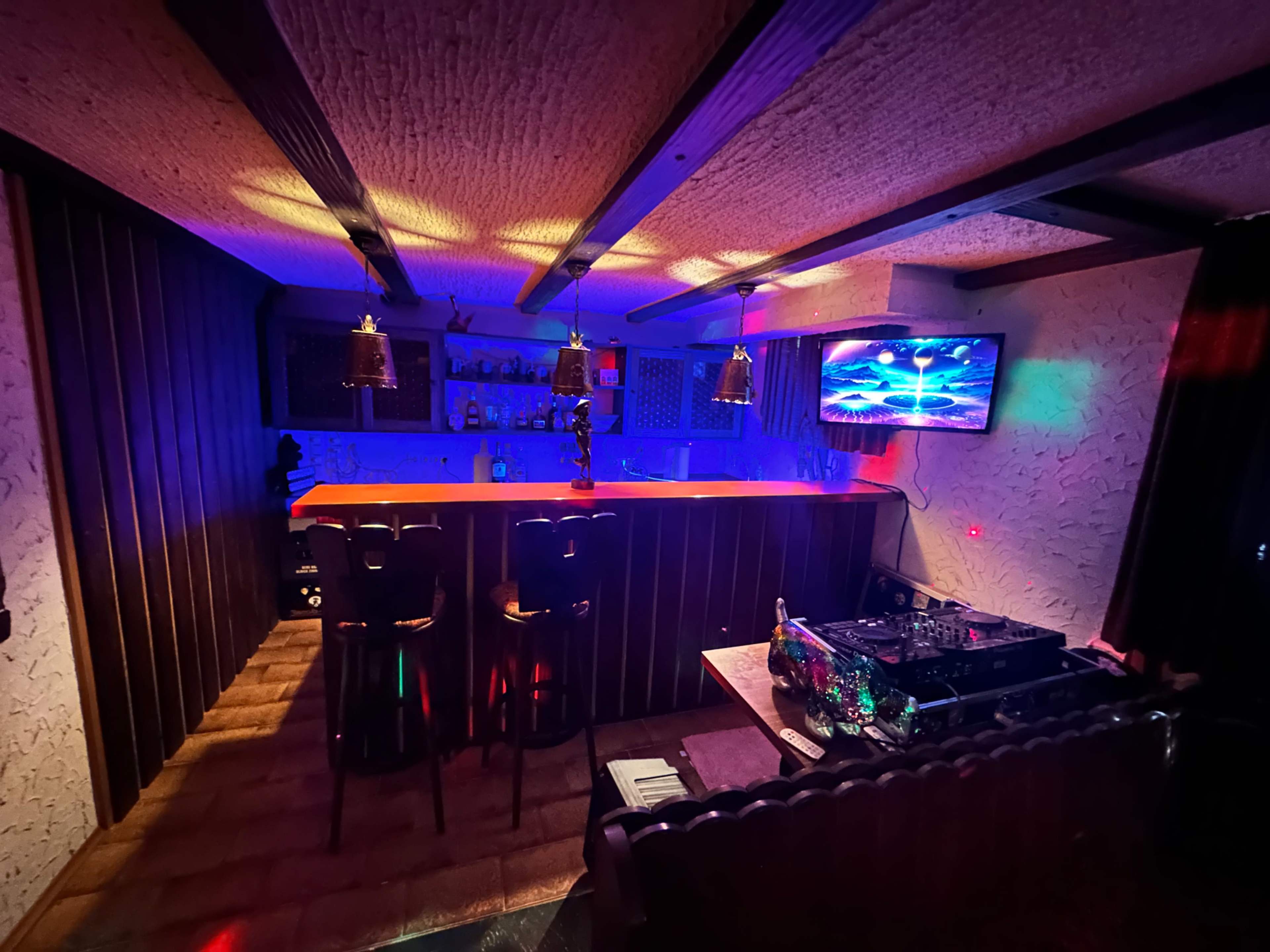 A dimly lit bar area featuring a counter with stools, a DJ setup, and a television displaying colorful visuals.