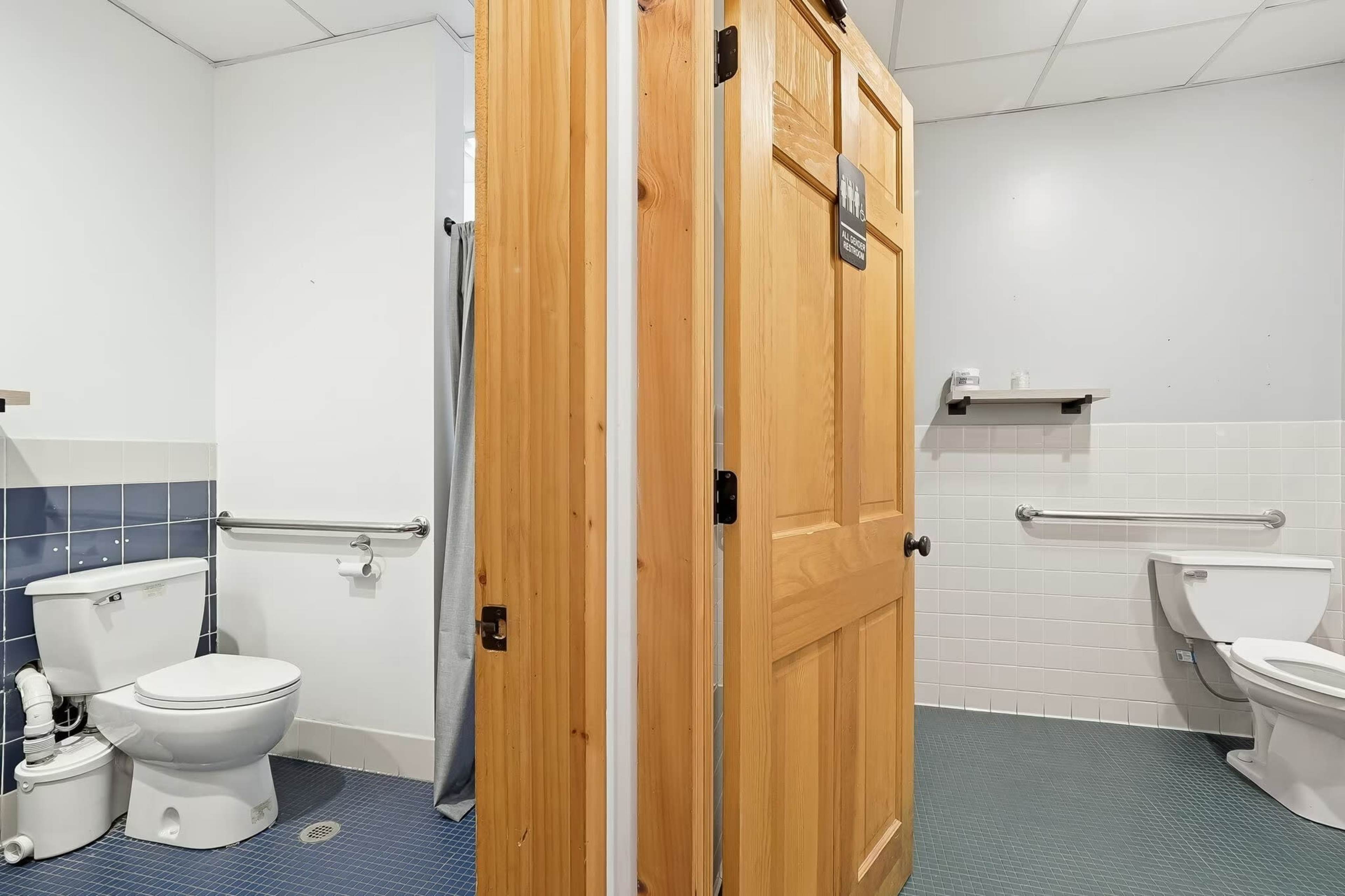 The image shows a restroom with two stalls separated by a wooden door, featuring a toilet on either side and tiled walls.