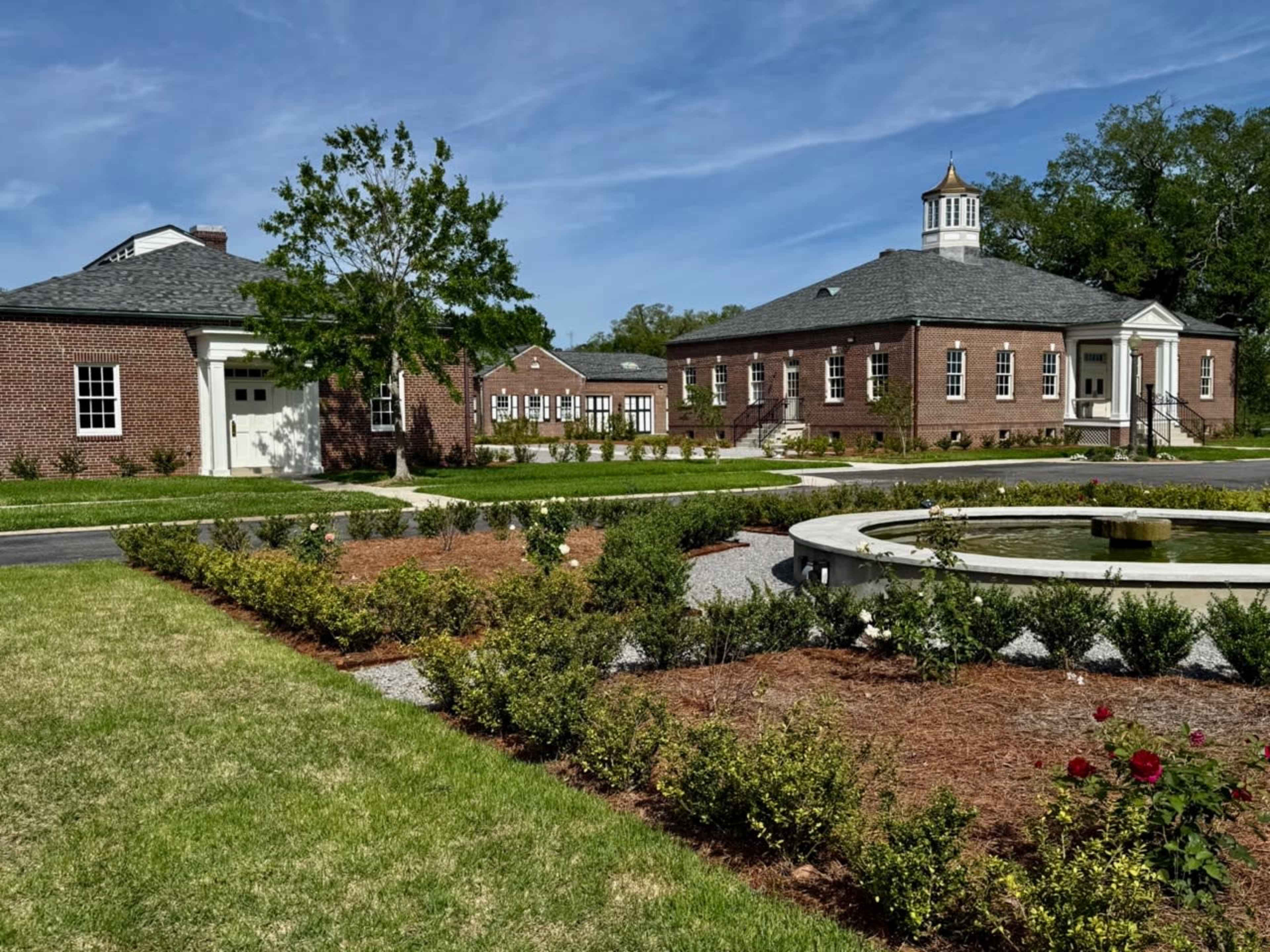 The image shows a landscaped area with a fountain, surrounded by brick buildings and greenery under a clear blue sky.