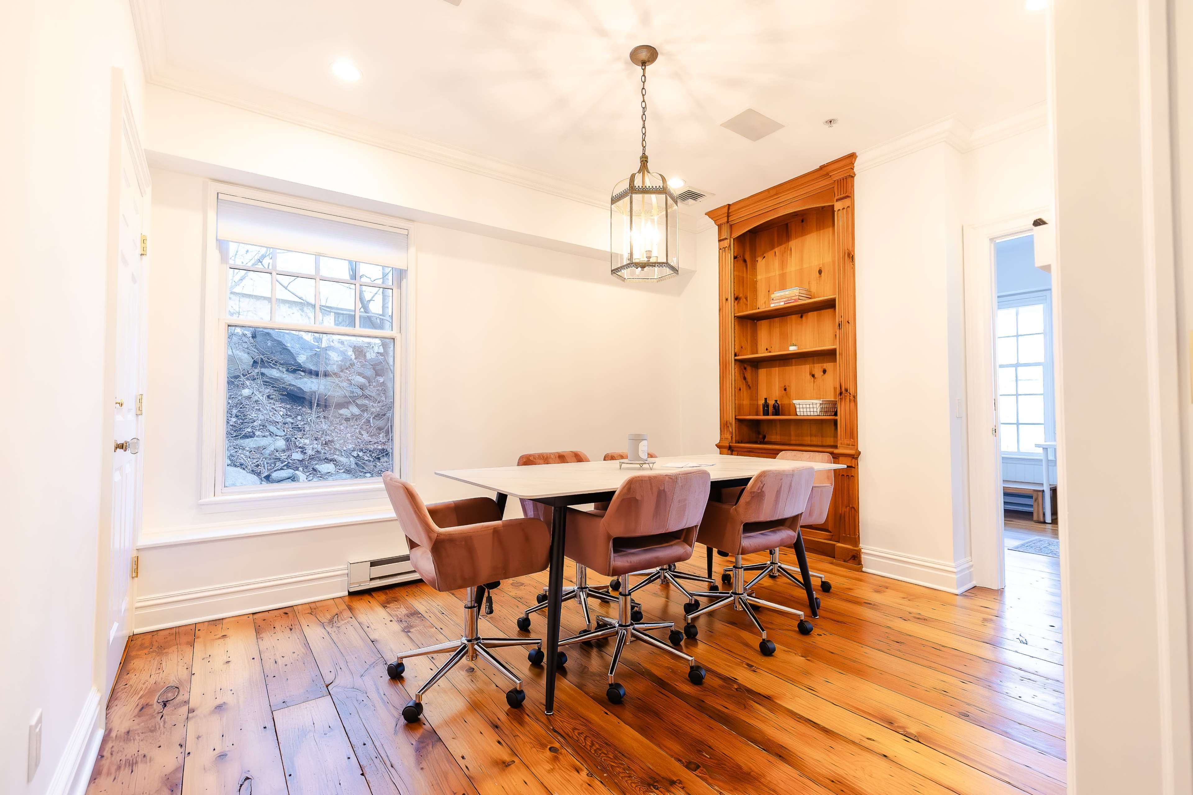 A conference room features a wooden table surrounded by rolling chairs, with a large window and a built-in wooden shelf against the wall.
