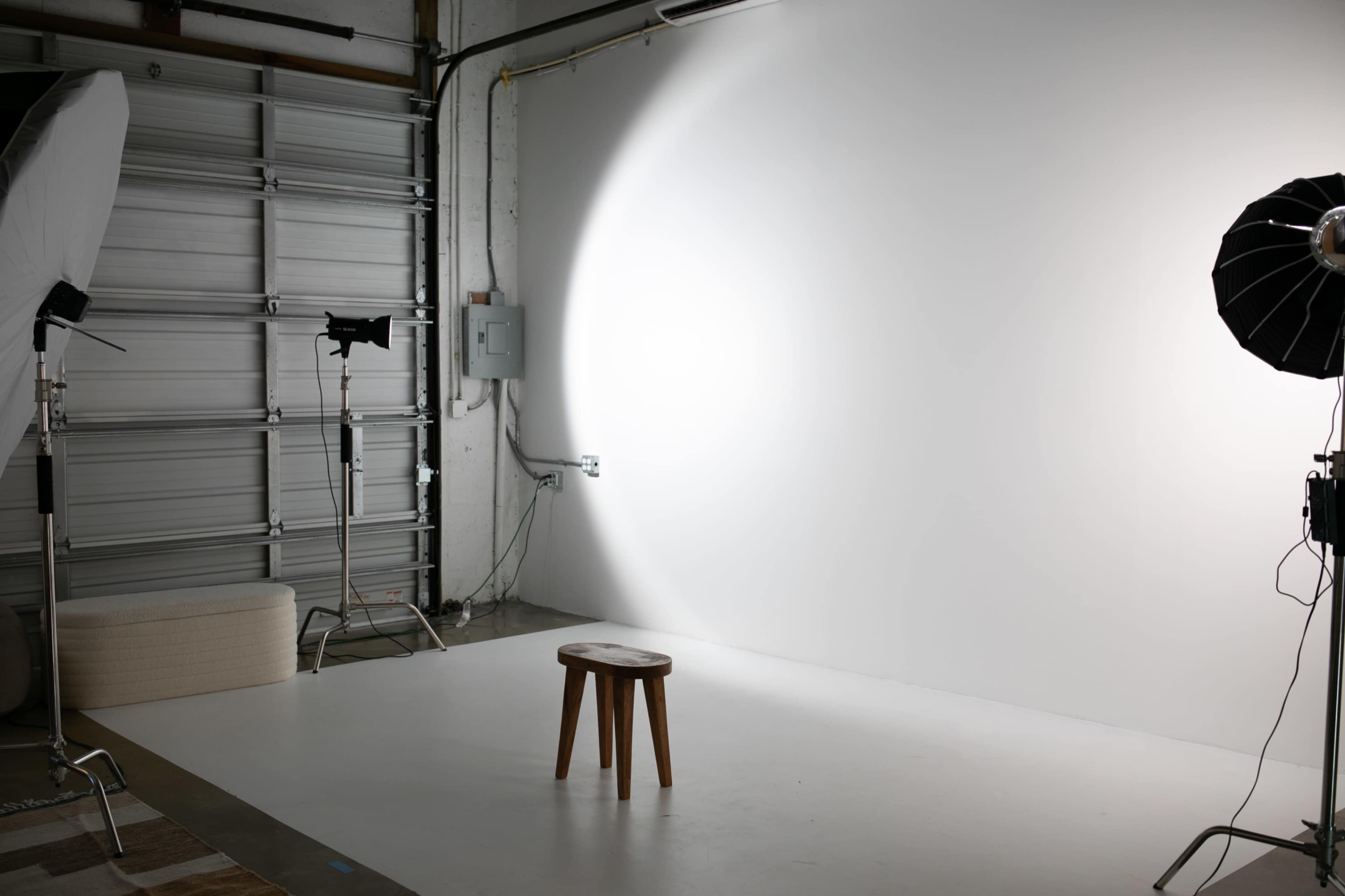 The image shows a photography studio with a wooden stool in front of a plain white backdrop, illuminated by studio lights.