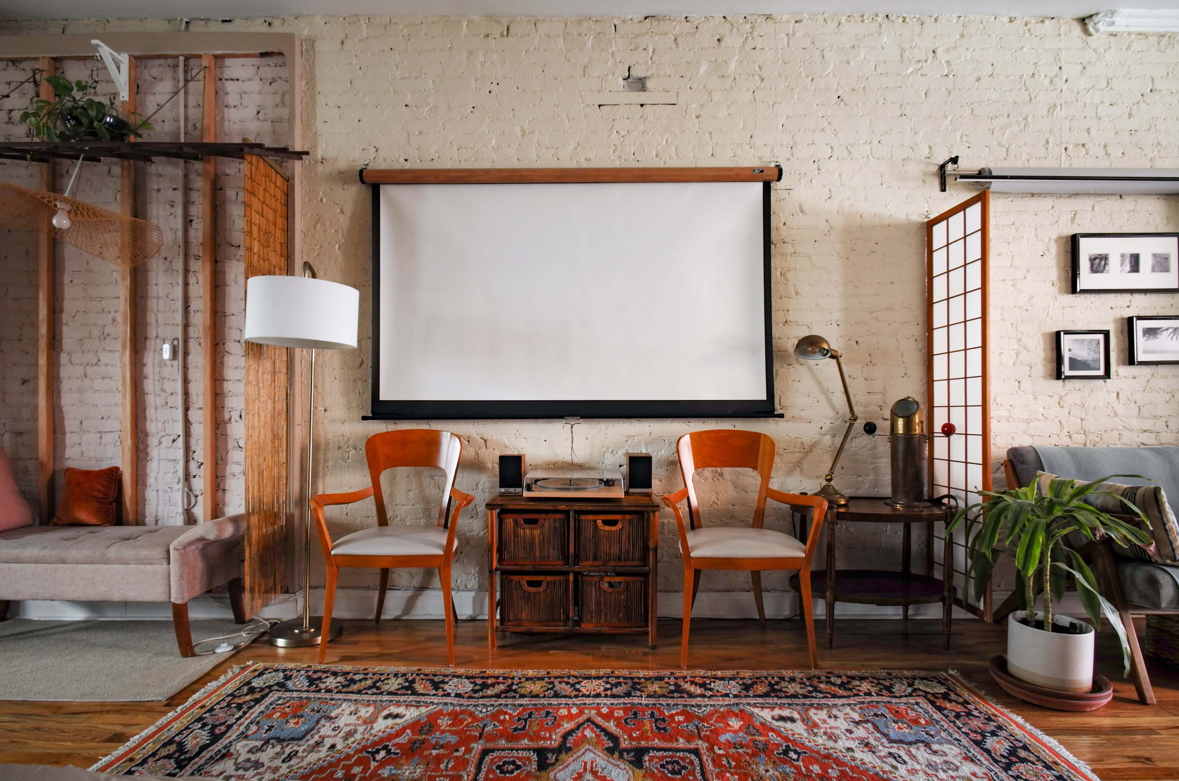 A cozy living room features two wooden chairs facing a blank projection screen, with a patterned rug and various decorative items along the walls.