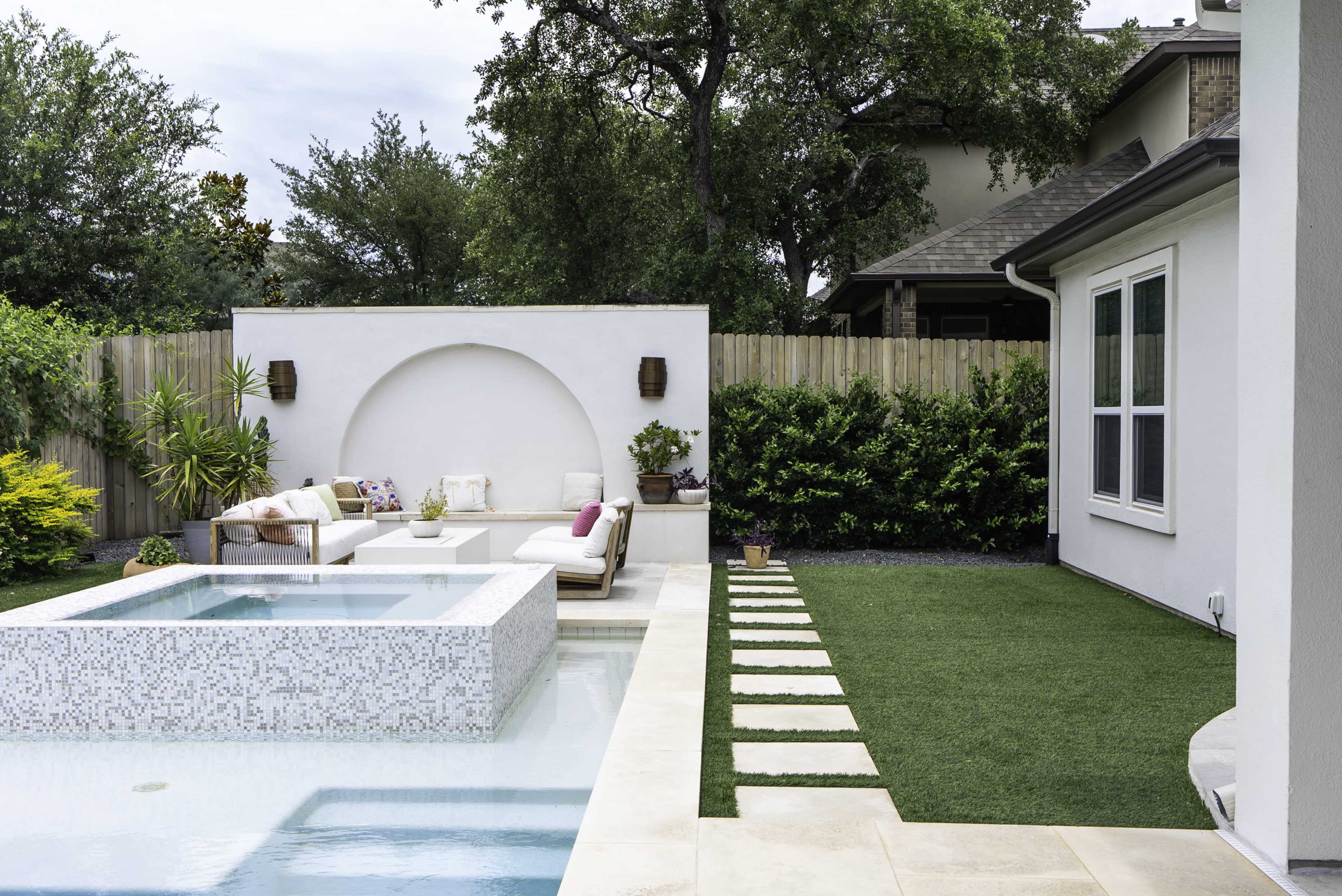 The image shows a modern backyard with a rectangular pool, a tiled spa, a seating area with cushions, and a neatly paved pathway alongside well-maintained greenery.