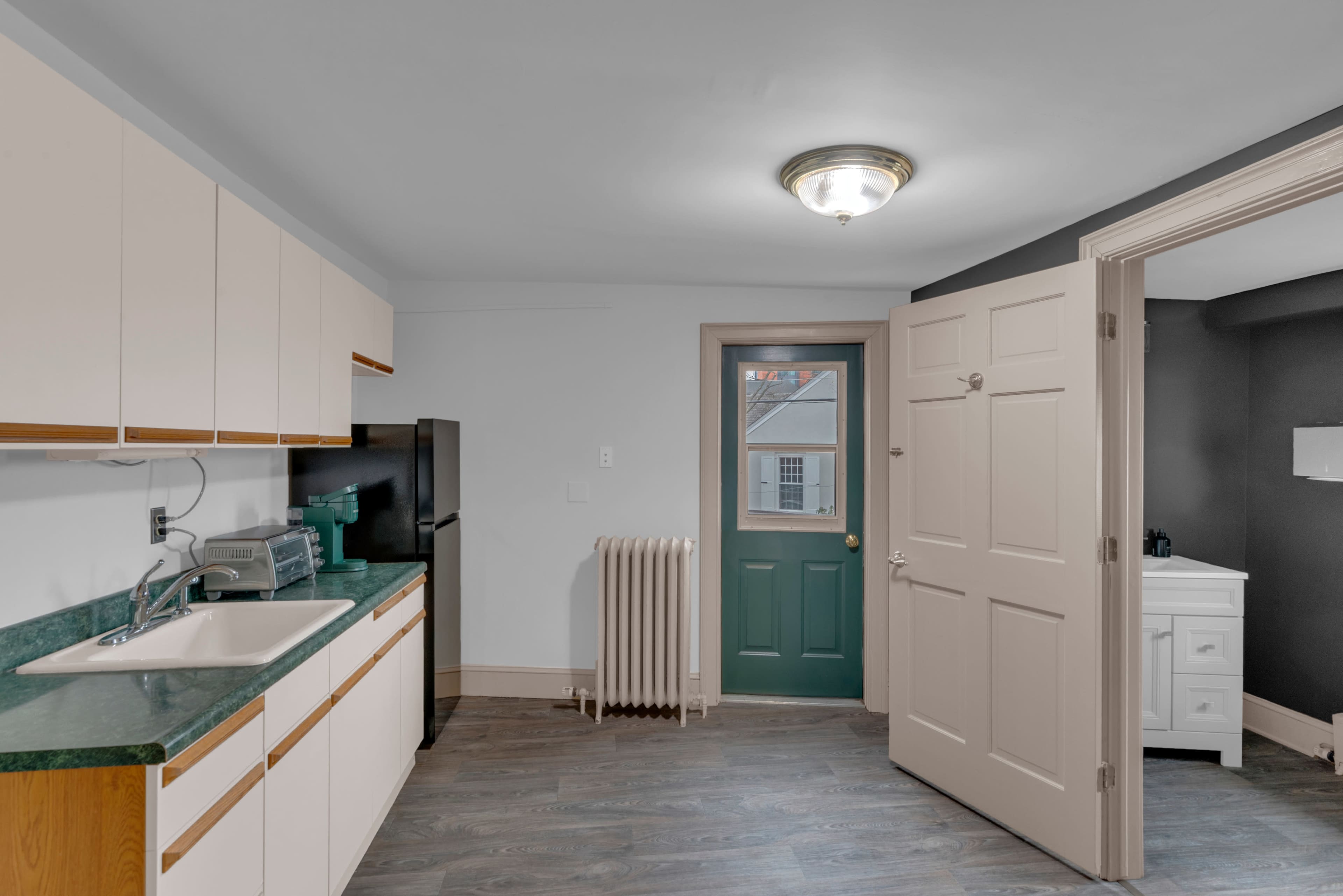 The image shows a kitchen area with a sink, refrigerator, and countertops, adjacent to a door leading outside and a door to another room.