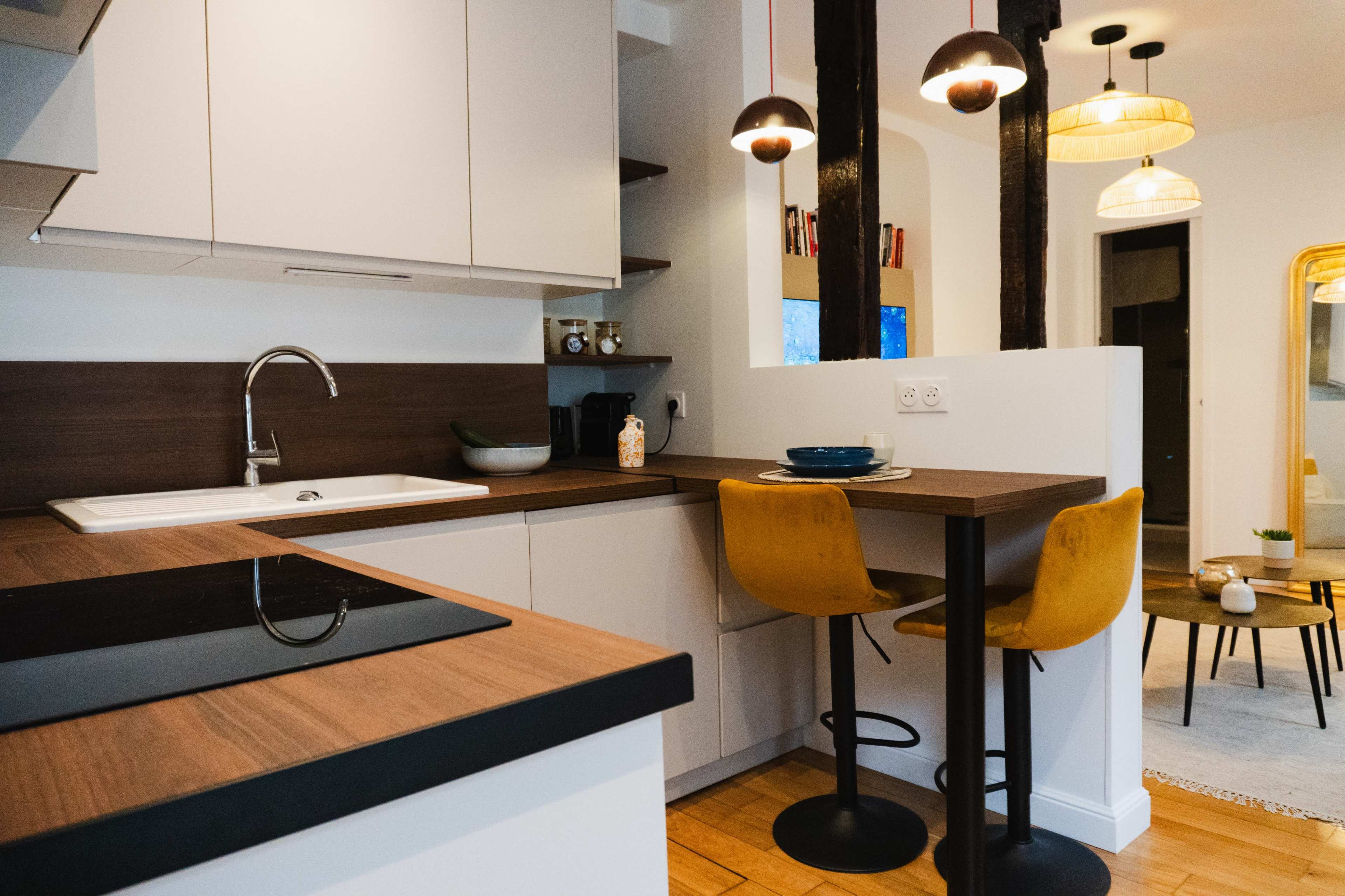 The image shows a modern kitchen featuring a sleek countertop, a sink, and two yellow bar stools adjacent to a small dining area.