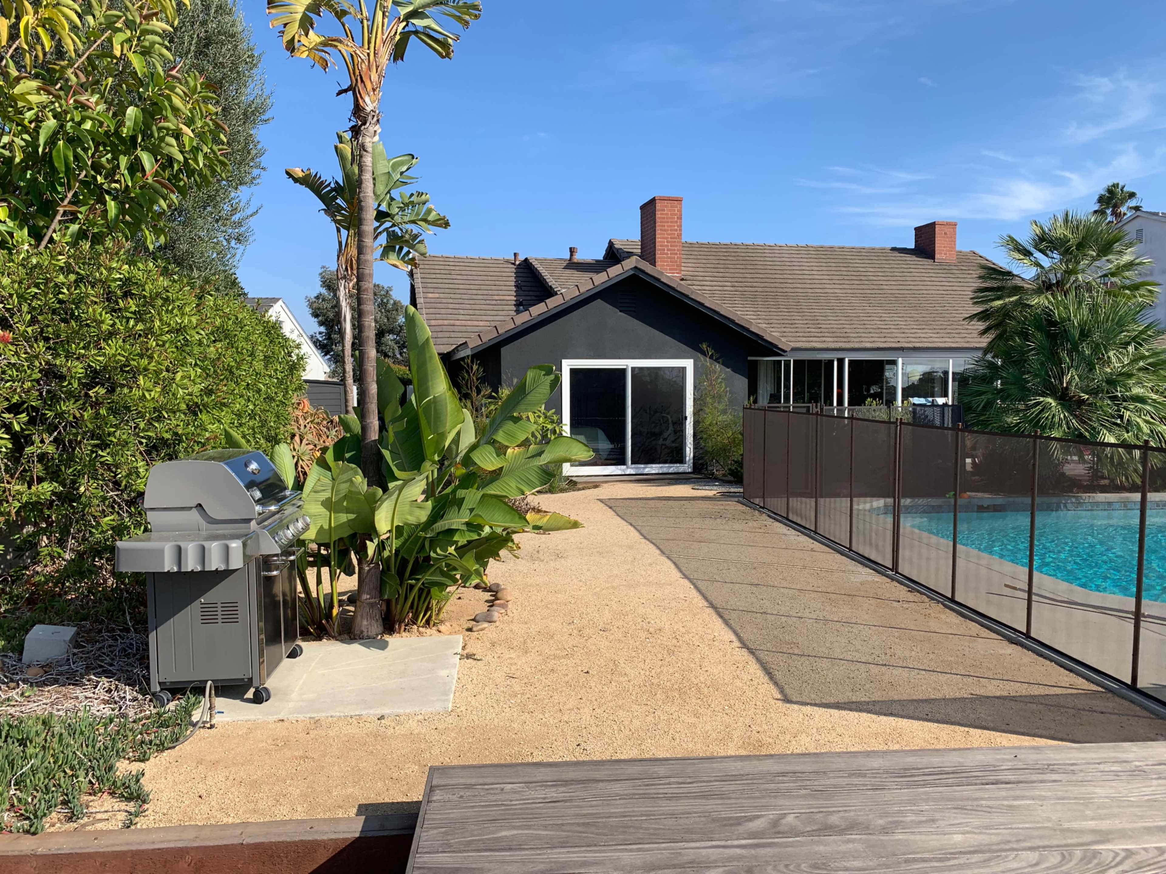 A backyard with a swimming pool, a barbecue grill on the left, and a house in the background surrounded by greenery.