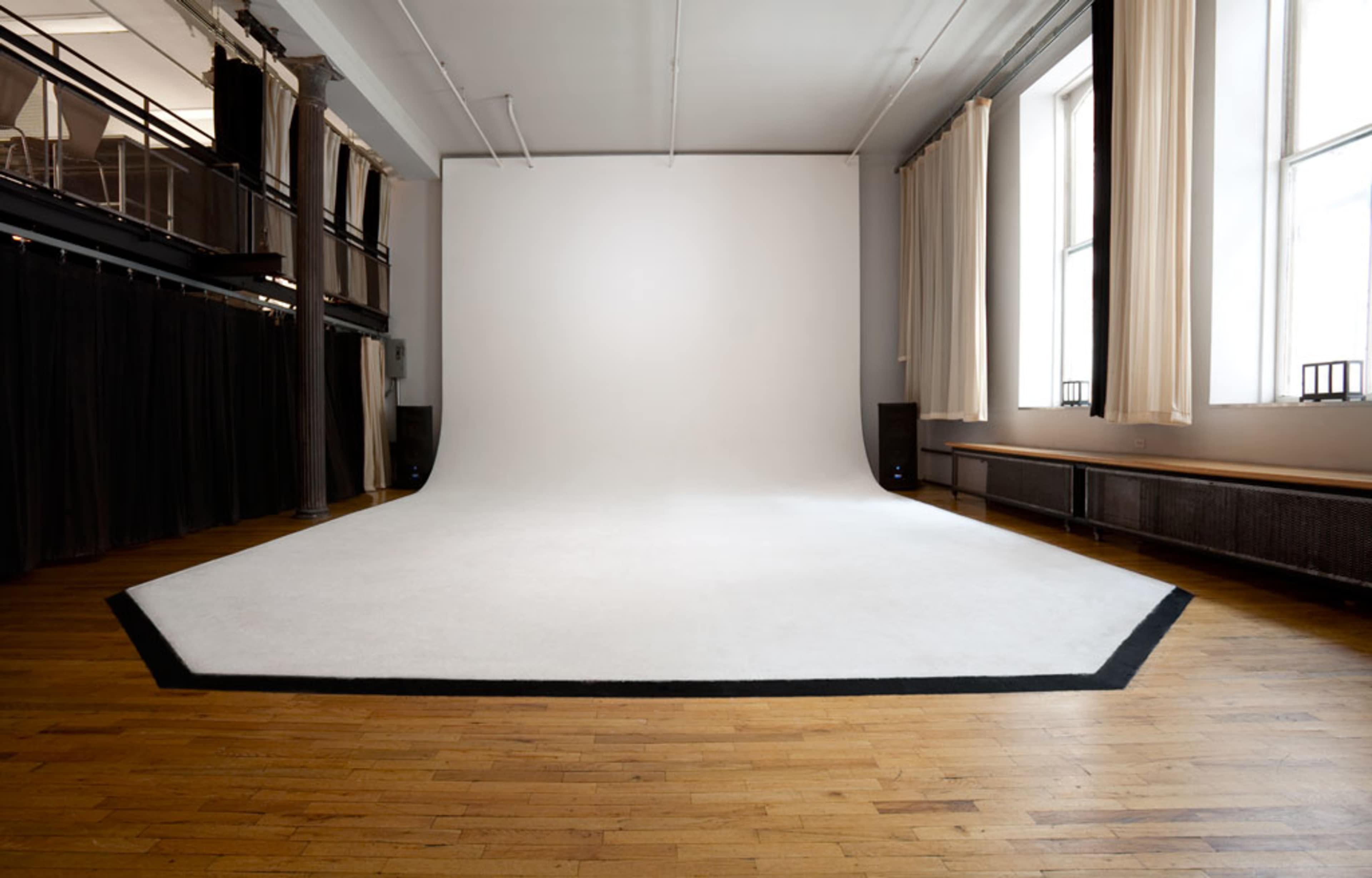 The image shows a spacious photo studio with a large, white backdrop and a wooden floor, illuminated by natural light from windows.