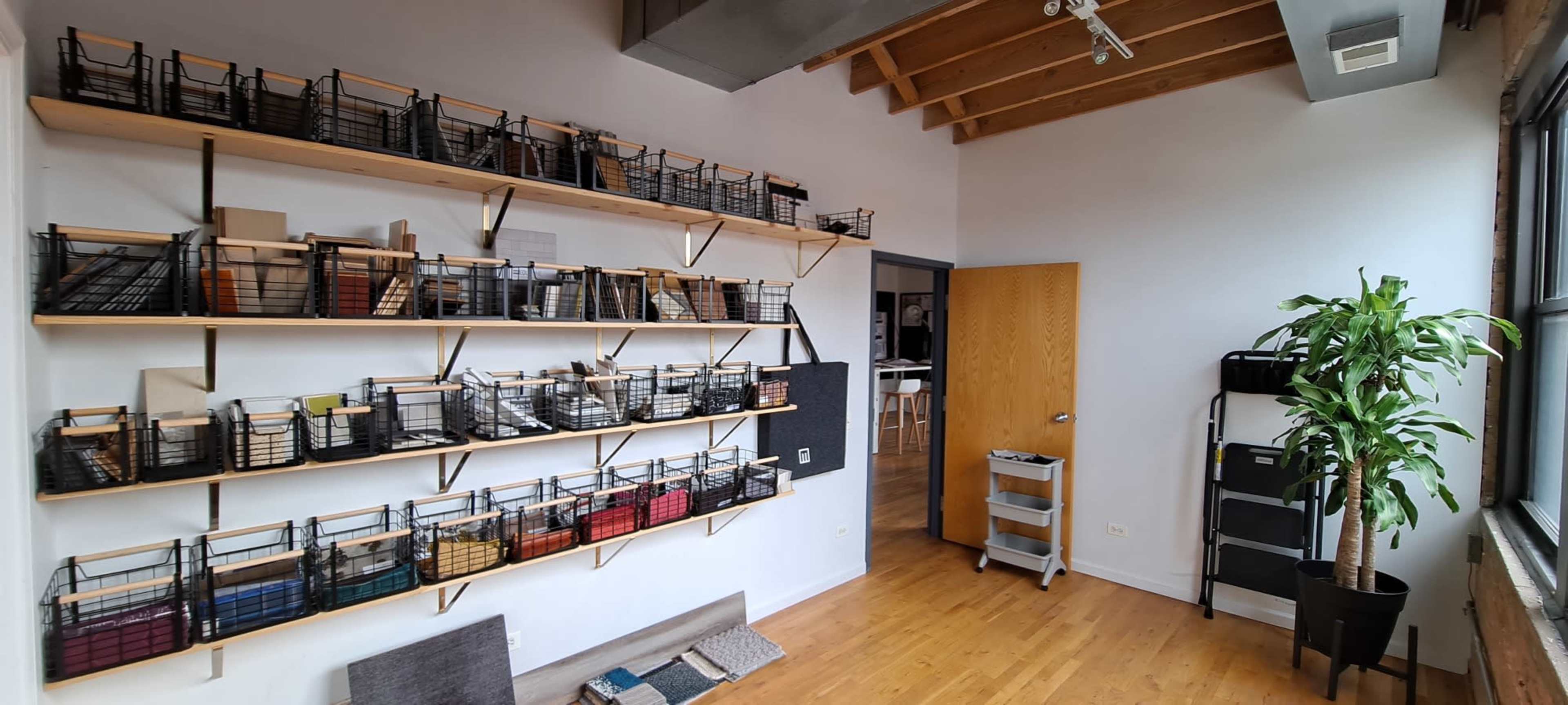 A room with wooden floors and a wall lined with shelves holding black wire baskets filled with various items, alongside a door and a plant in the corner.