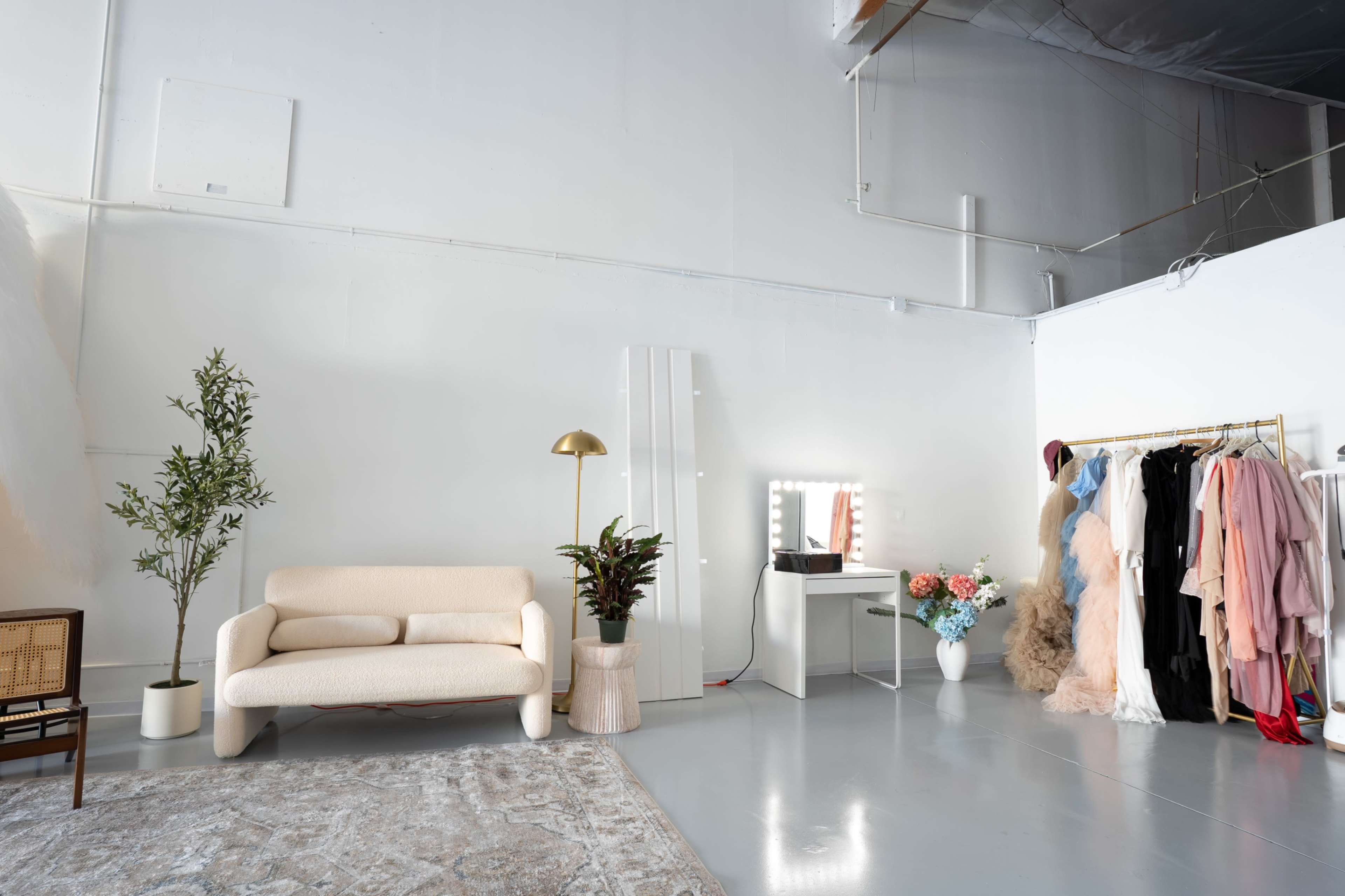The image shows a bright, minimalistic studio space featuring a beige sofa, a tall plant, a vanity mirror with lights, and a clothing rack filled with various garments.