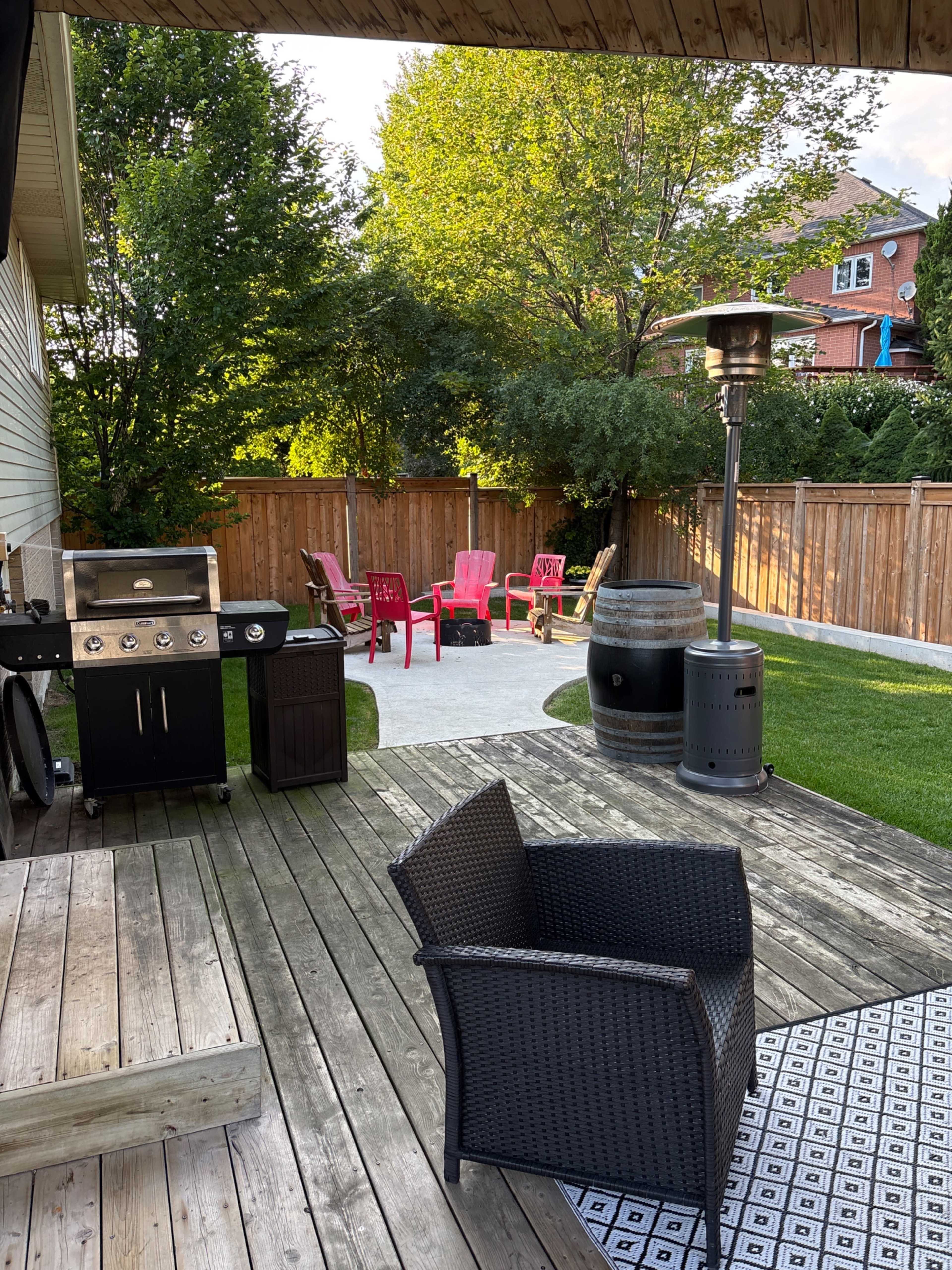 The image shows a backyard patio with a grill, a black wicker chair, a small table, and pink lawn chairs arranged around a fire pit, all surrounded by a wooden fence and greenery.