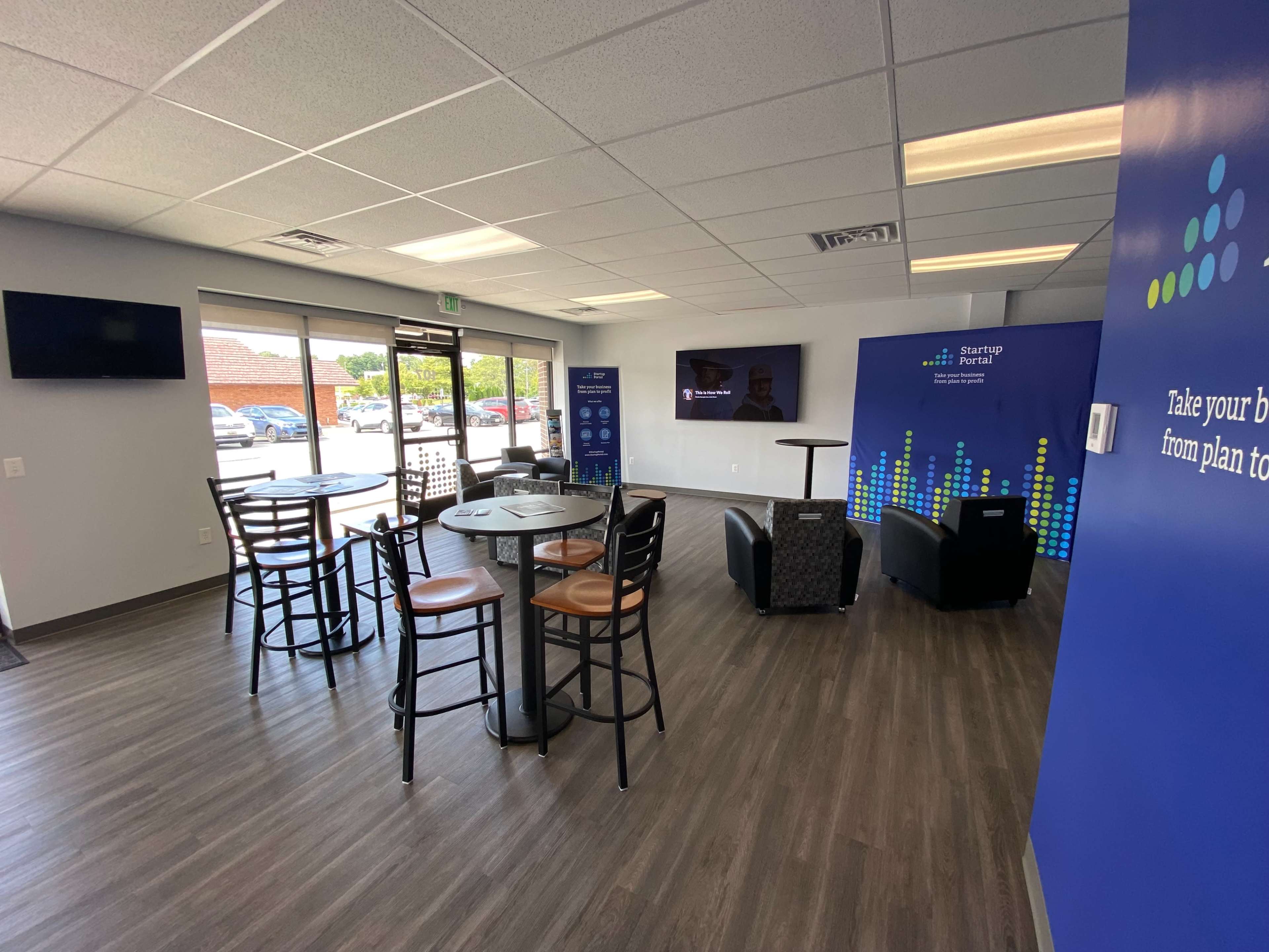 A modern office space with tables and chairs, a large screen, and windows allowing natural light.